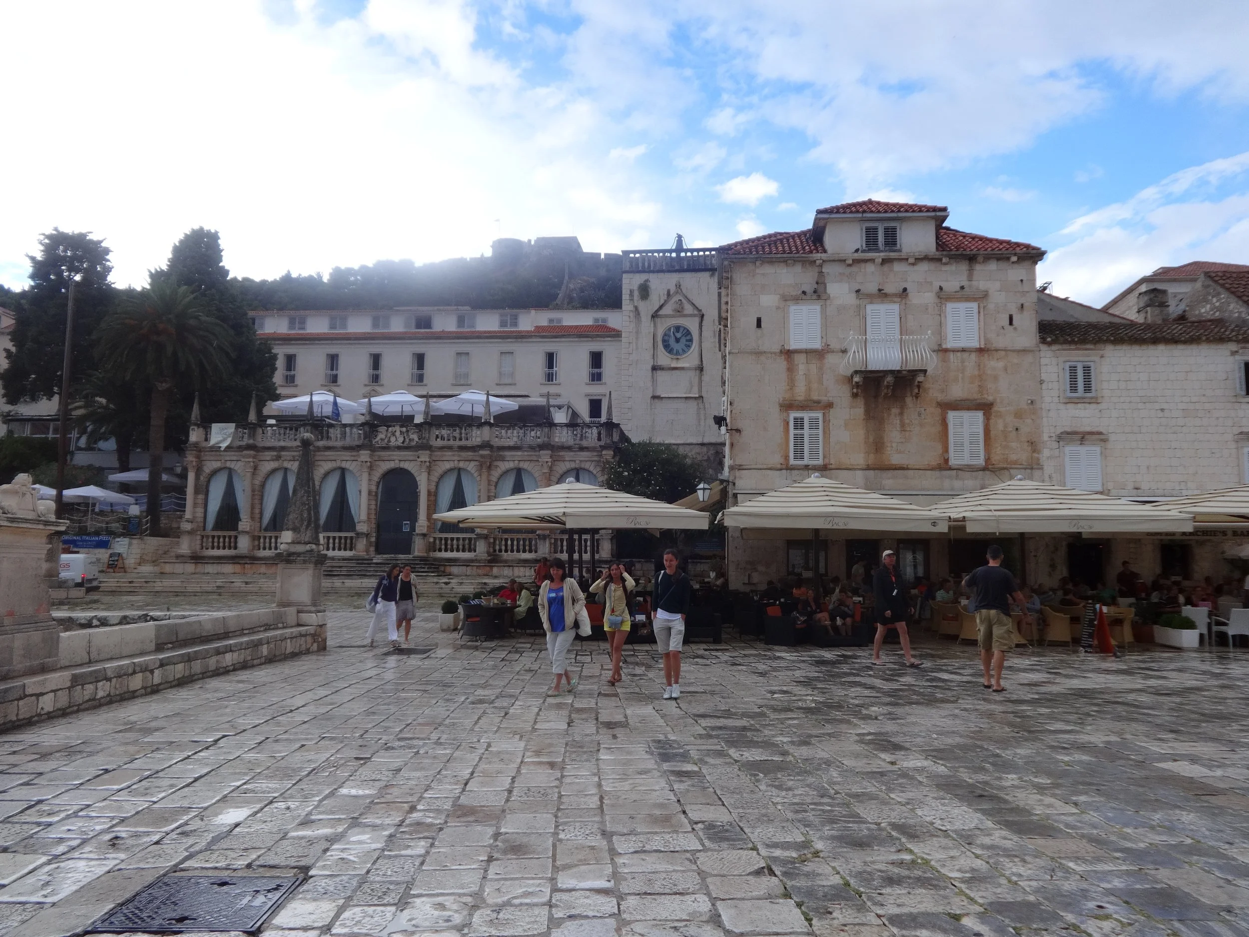 A lively outdoor square with people walking and sitting under umbrellas, historic buildings with white shutters, a clock tower, and a hillside castle in the background. The ground is old cobblestone.