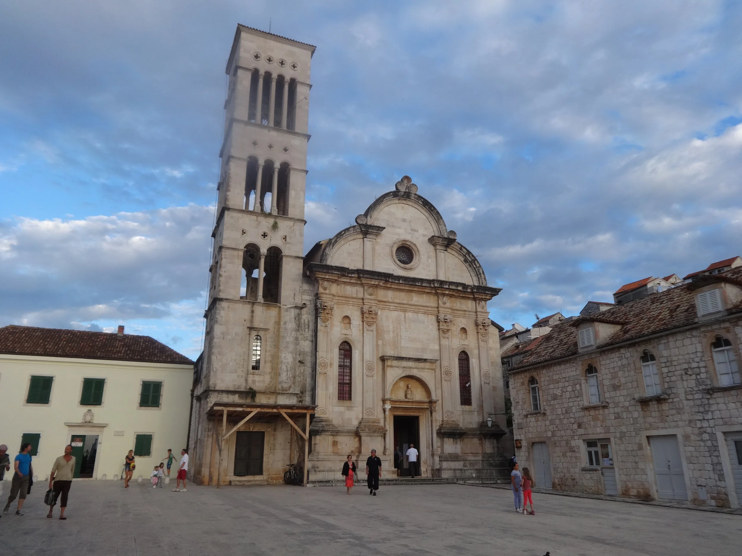 A historic church with a tall bell tower, located in a European town square, with people walking and gathering outside.