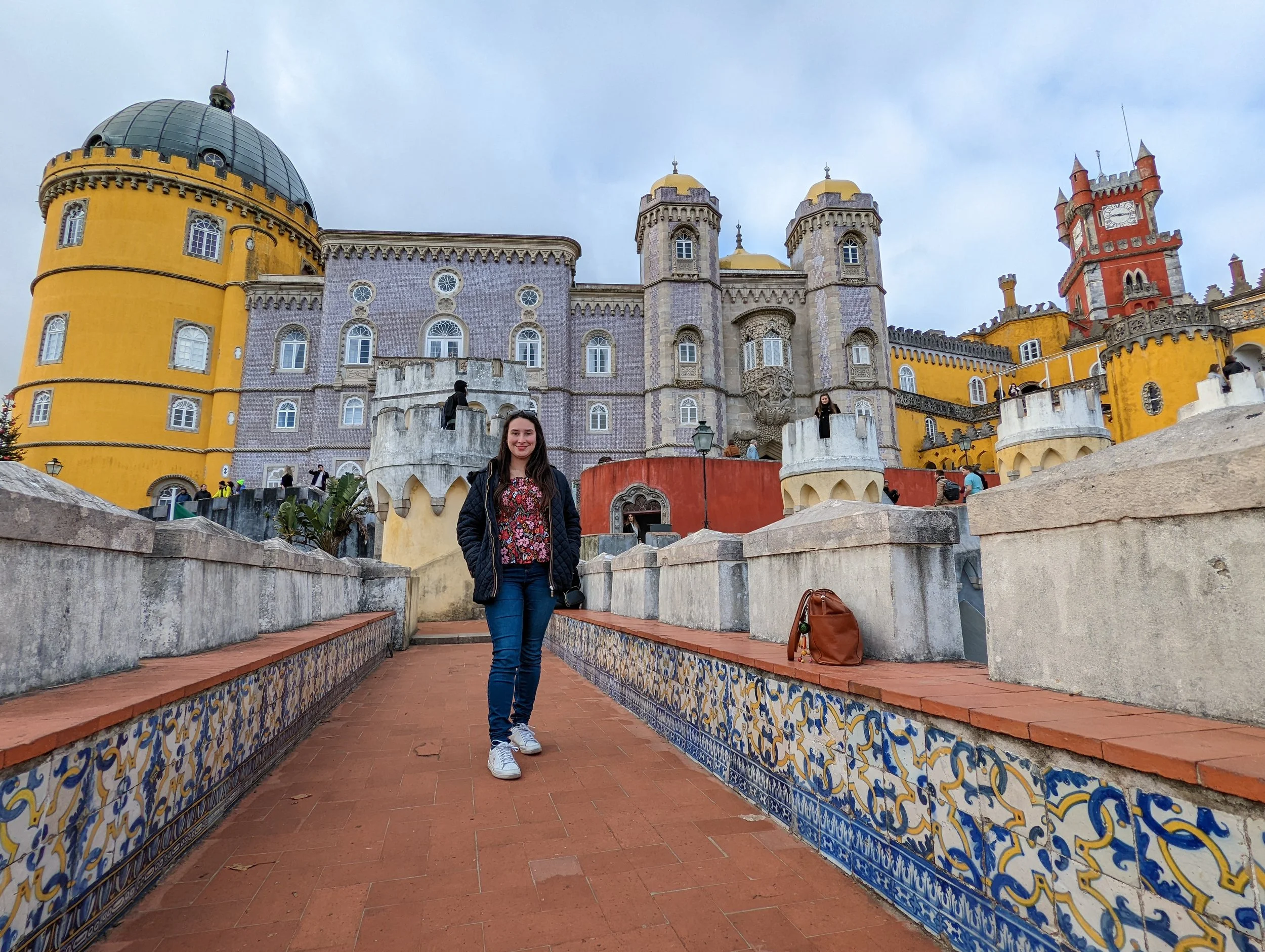 A woman with long dark hair, wearing a black jacket, floral shirt, blue jeans, and white sneakers, standing on a tiled pathway in front of colorful castle-like buildings with yellow, purple, and red turrets, under a cloudy sky. There are other visito