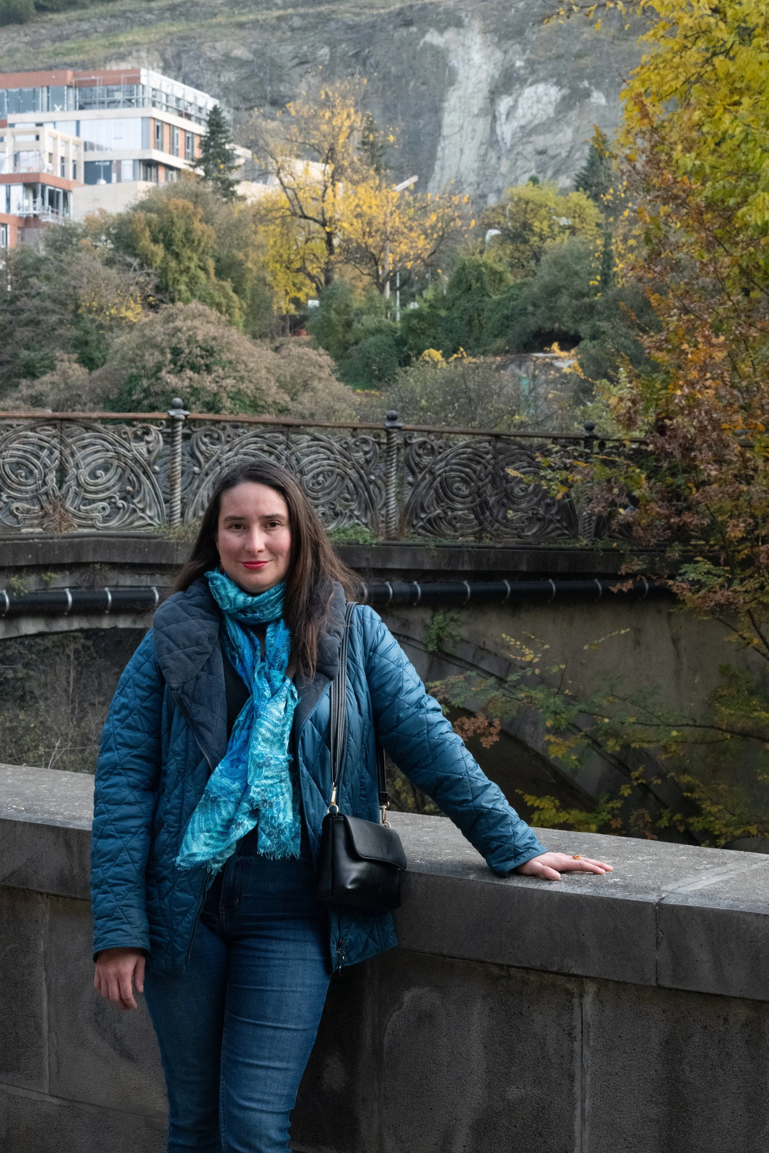 A woman with long dark hair and light skin, wearing a blue quilted jacket and a blue scarf, standing outdoors near a stone railing with her right hand resting on it. She is smiling with a backdrop of autumn-colored trees, a bridge, and buildings on a