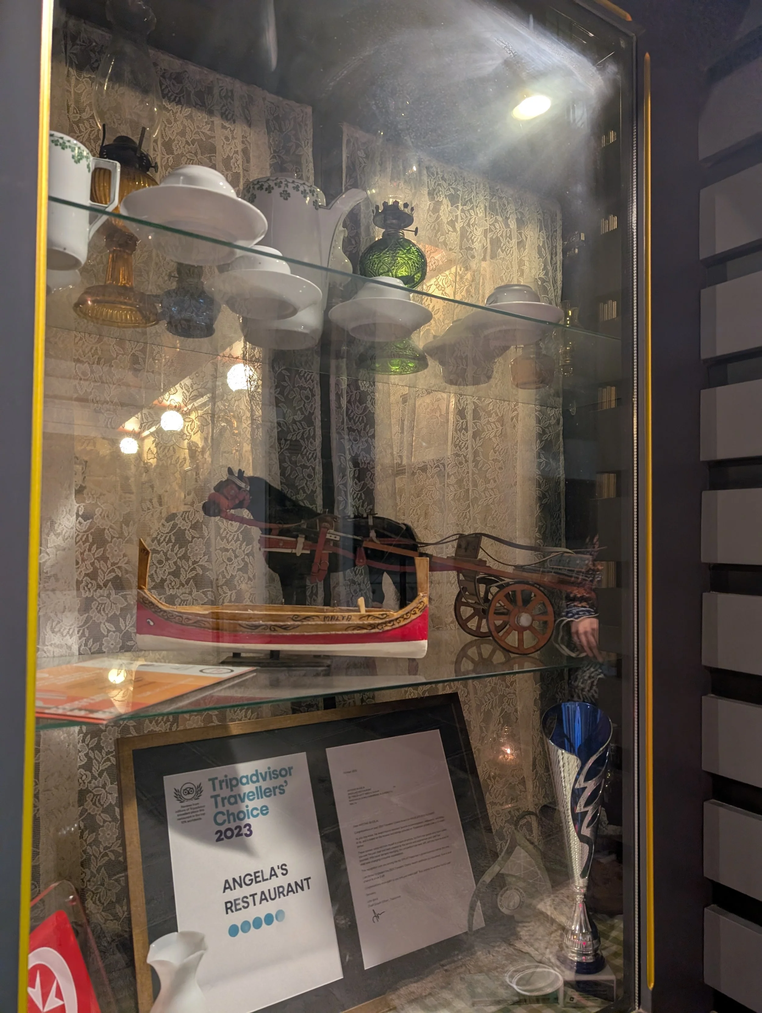 Display case with old-fashioned items including white dishes, a green glass lamp, a wooden wagon, a small sword, and a clear glass trophy. There is a sign for TripAdvisor awards and a document inside the case.