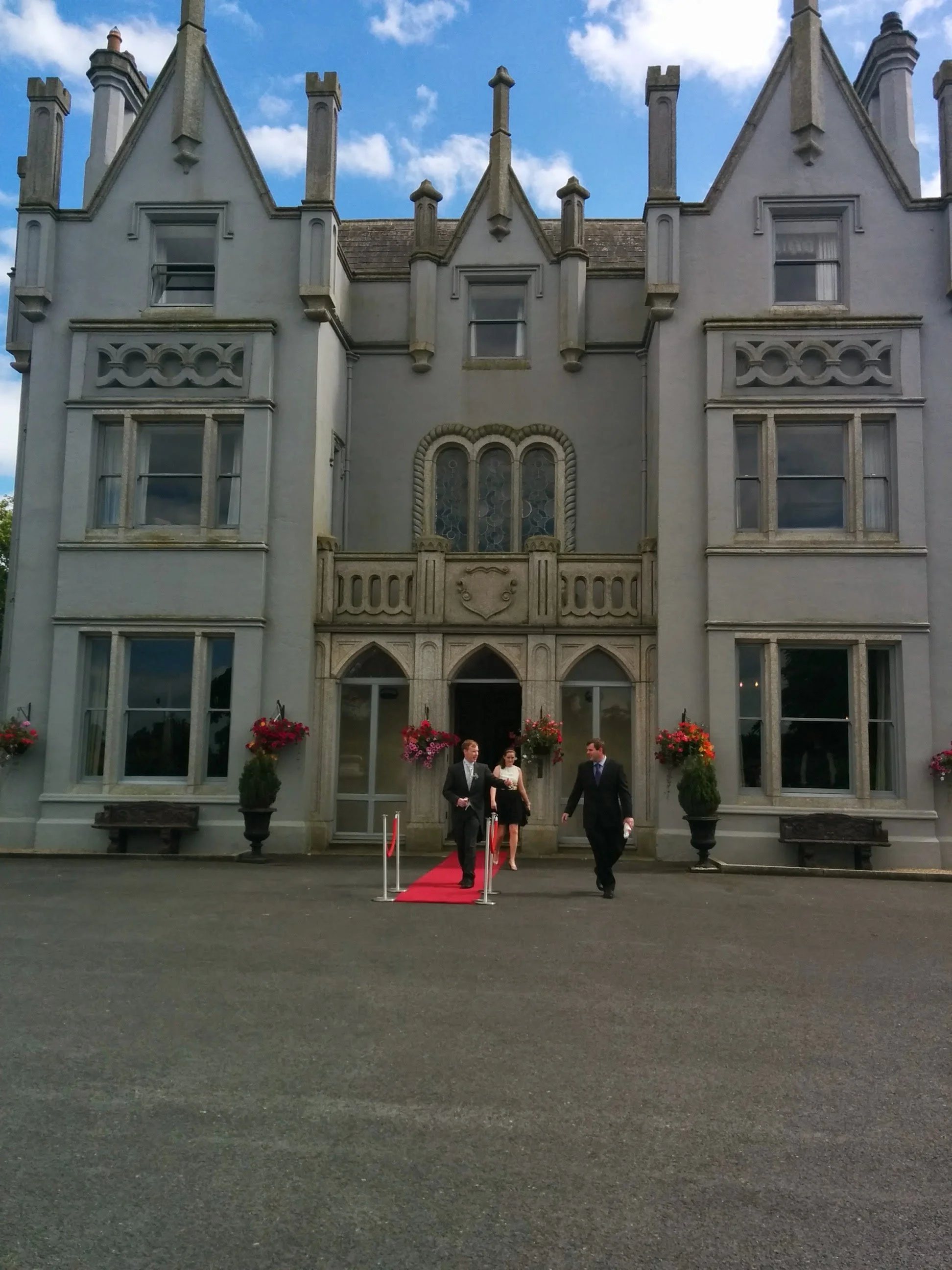 A large, ornate Victorian-style building with gray stone walls and a red carpet leading to the entrance. Three people, two men in suits and a woman in a dress, walk down the red carpet, which is flanked by potted flowers. The building has multiple wi