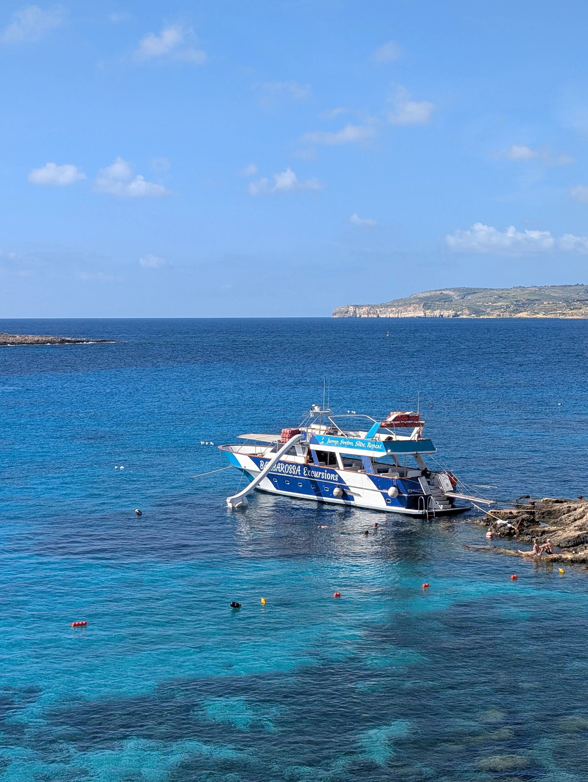 A boat named BELLAROSSA Excursions anchored near a rocky coastline with clear blue water and a distant hillside with cliffs. Swimmers are in the water.