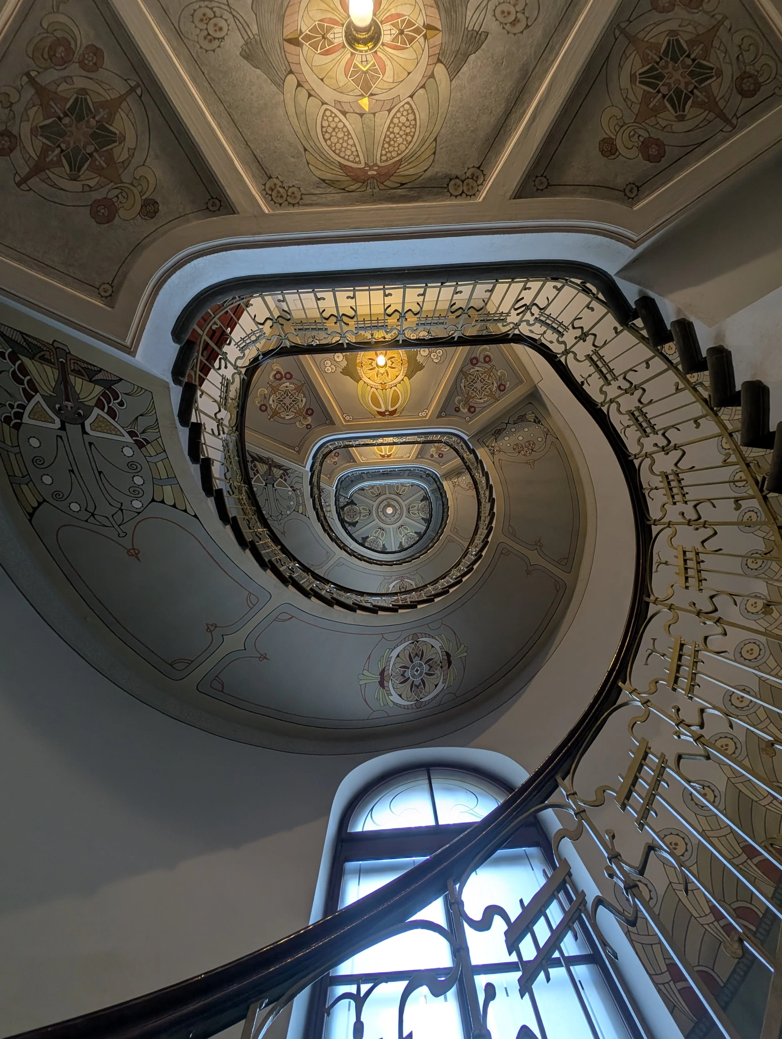 Decorative staircase interior with ornate, painted ceiling designs, and a spiral view looking up from the bottom of the staircase.