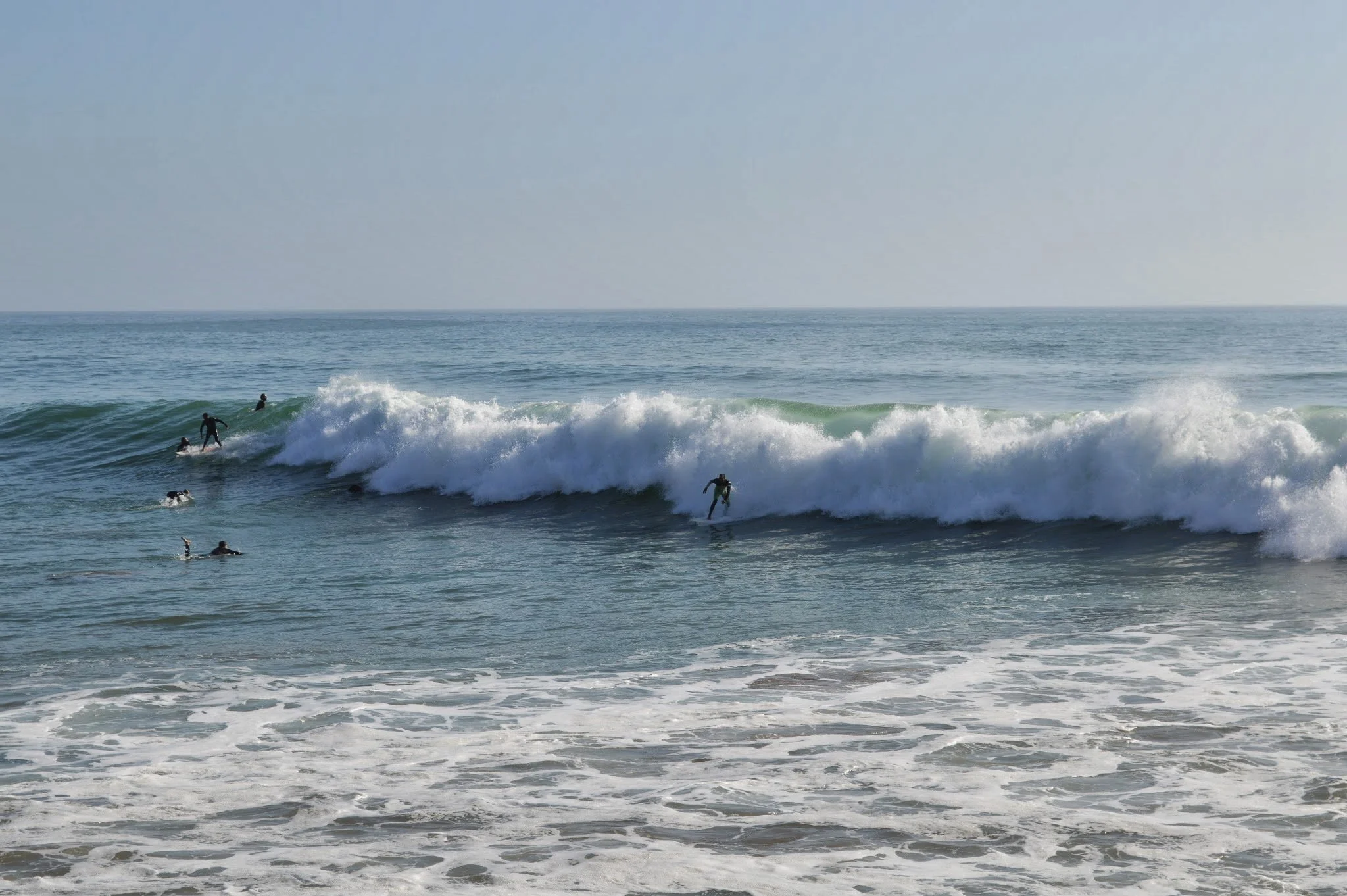 People surfing and swimming in the ocean with waves breaking near the shore, under a clear sky.