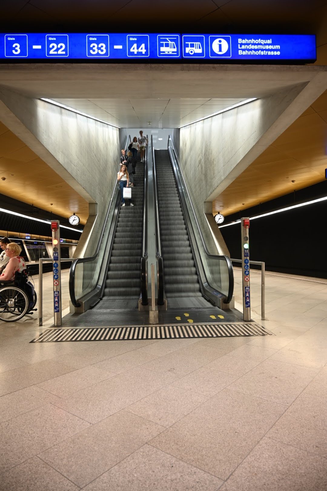 Inside a modern train station with an escalator heading up, passengers, including a person in a wheelchair, waiting near the escalator, with a blue digital sign overhead showing train platform information and an information icon.