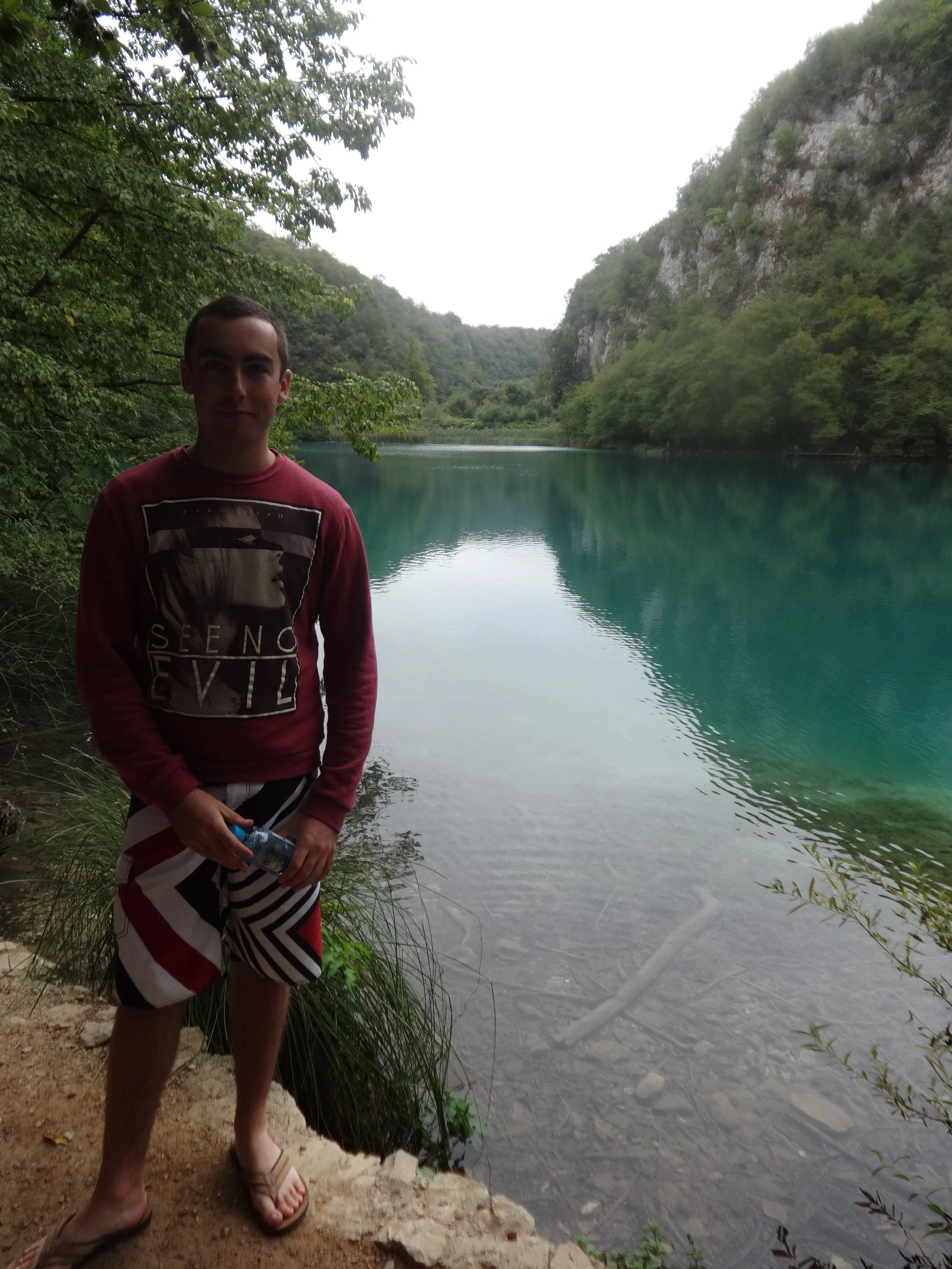 A man standing on a dirt and rock shoreline next to a calm, greenish-blue body of water in a forested area with hills or cliffs covered in trees.