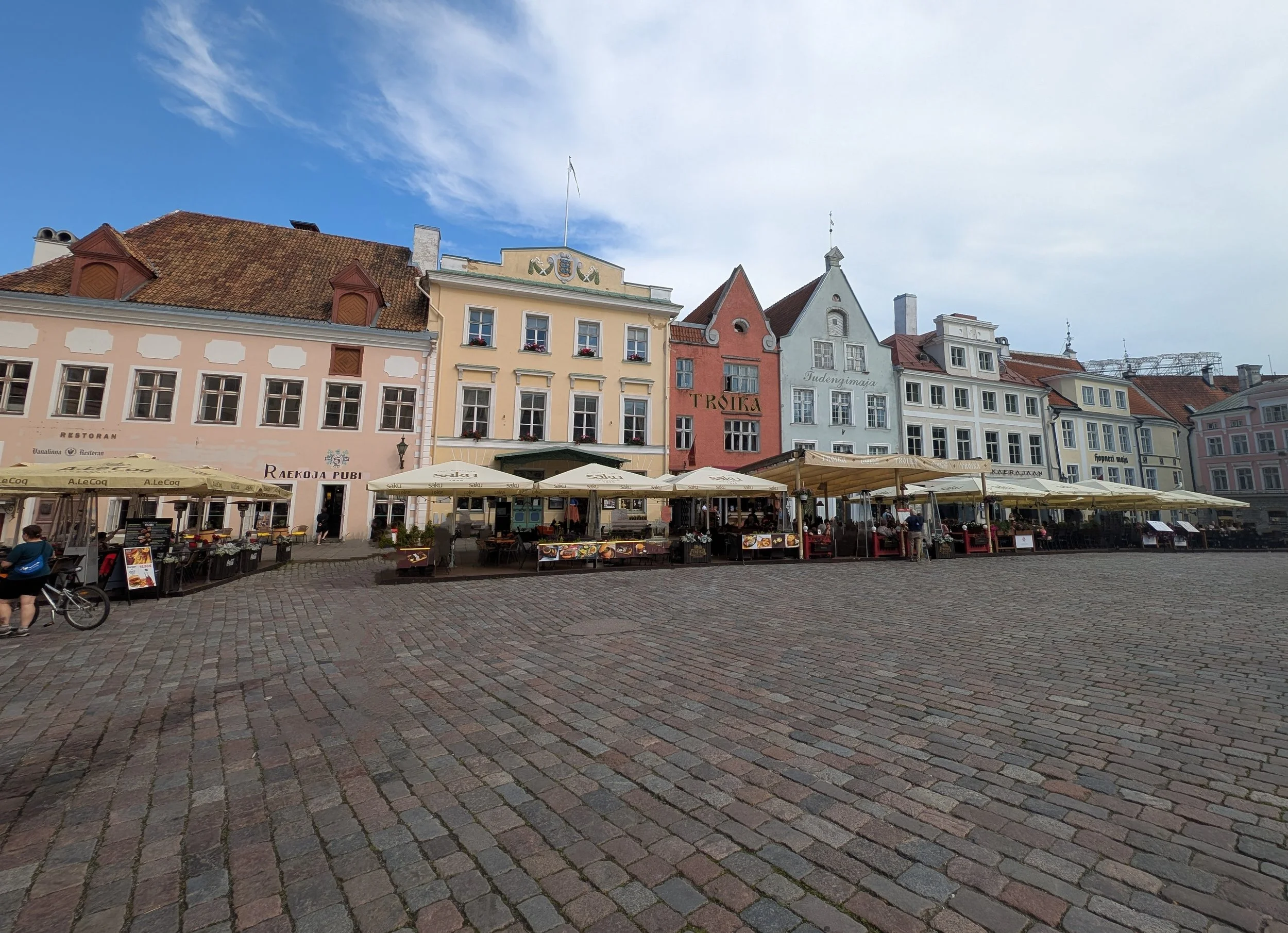 A town square with colorful historic buildings, outdoor cafes with umbrellas, cobblestone pavement, and a partly cloudy sky.
