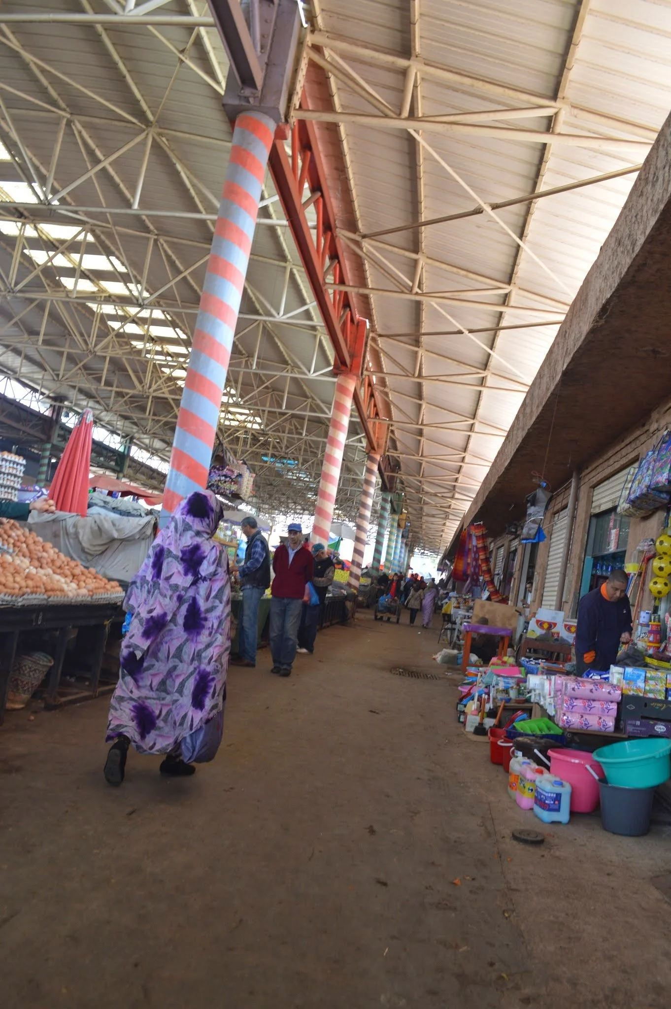 View of a busy indoor market with vendors selling various goods, including eggs and household items, under a high, open ceiling with a mix of metal and wooden structures.