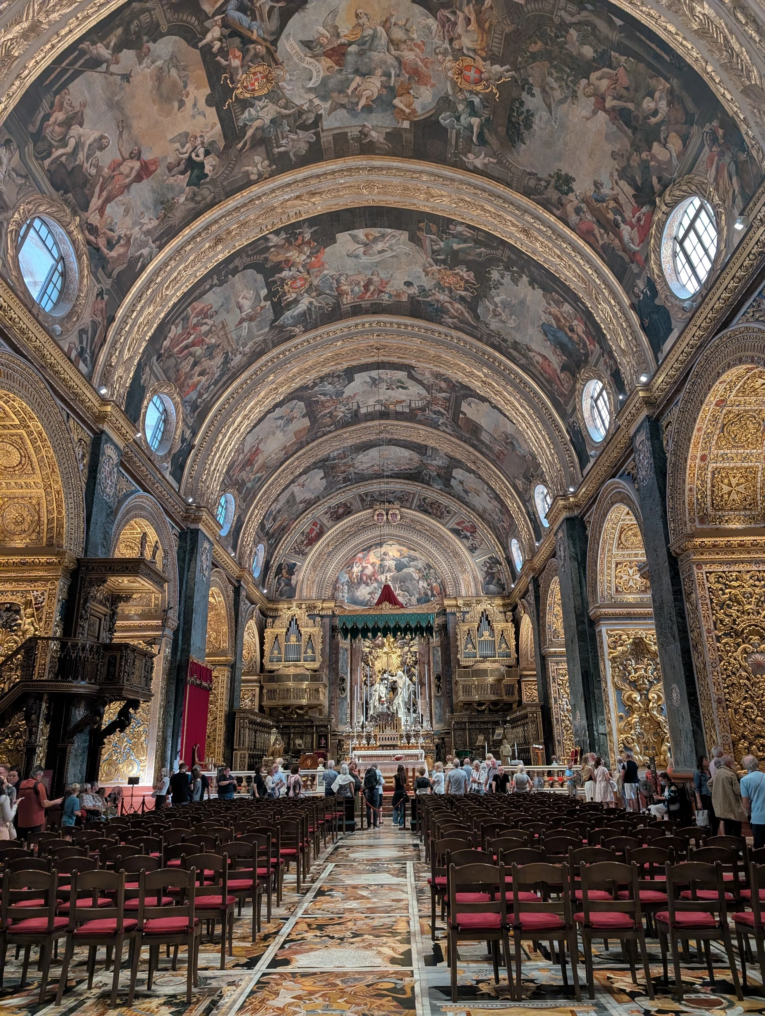 Interior of a richly decorated church with elaborate gold detailing, painted domed ceilings, and a congregation of people gathered near the altar.