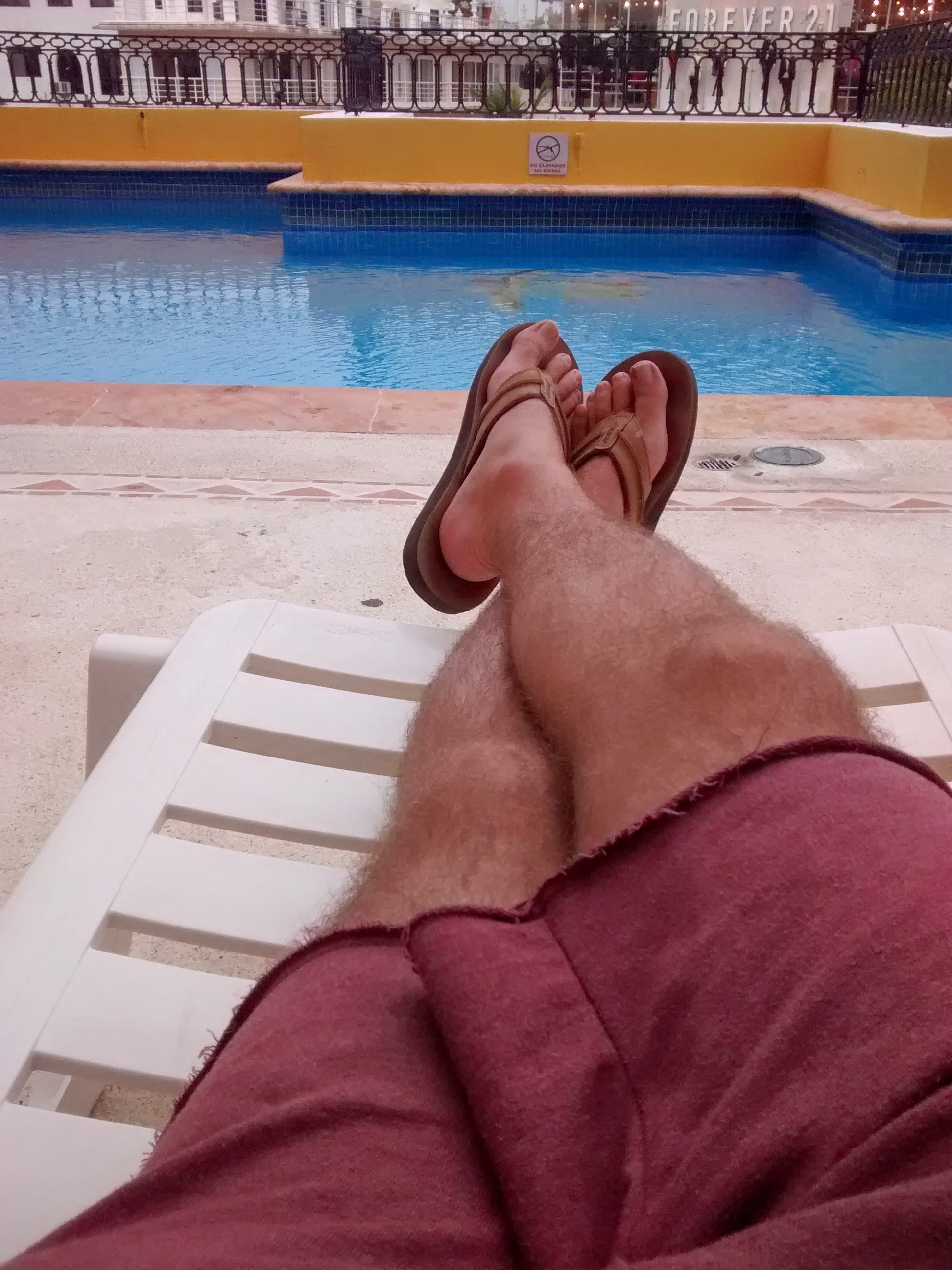 Person relaxing on a lounge chair with legs crossed, wearing sandals and a red shirt, near a swimming pool in an outdoor area during daytime.