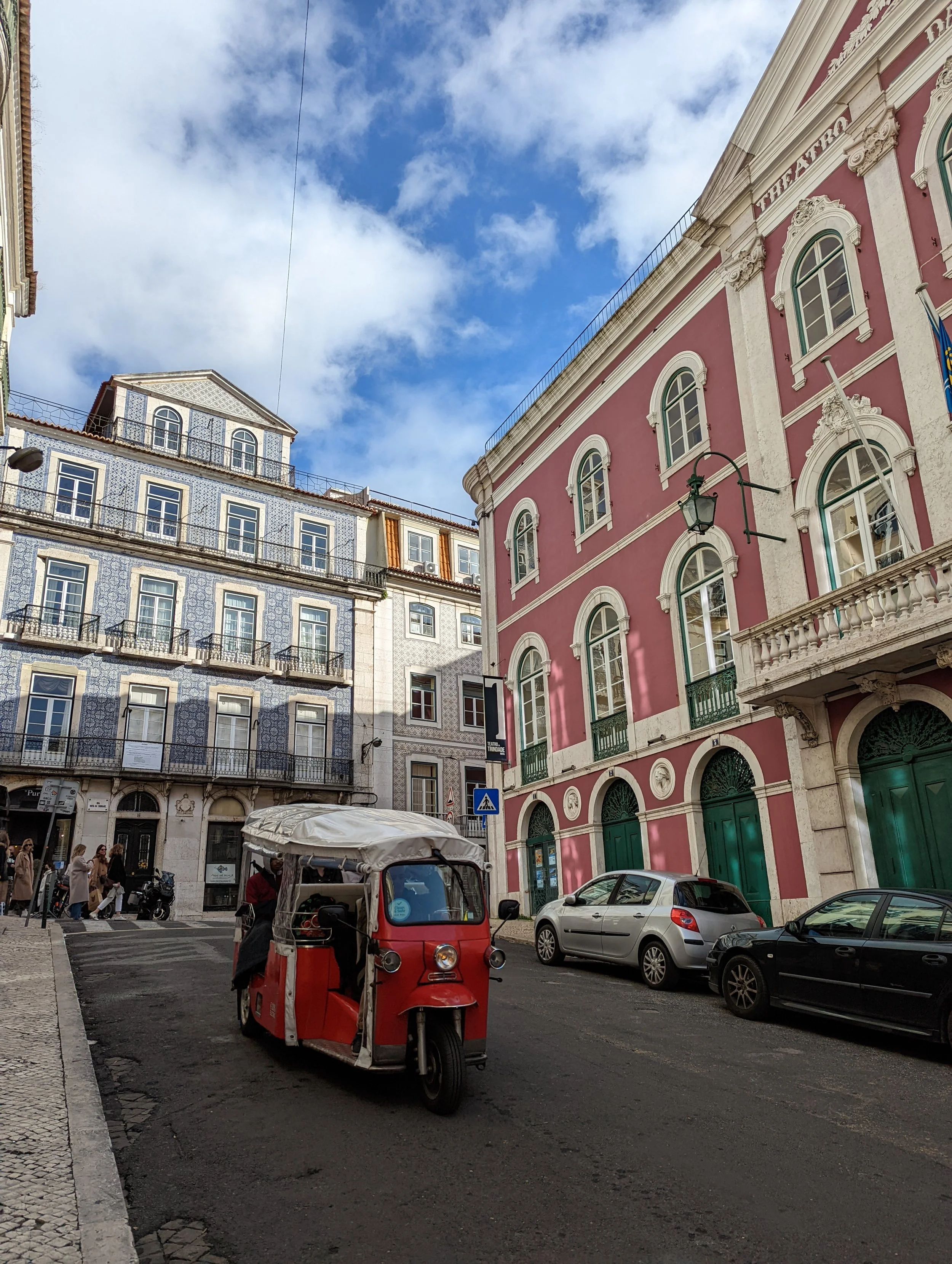 A street scene with a pink and white ornate building, a blue and white tiled building, cars parked along the street, a red three-wheeled motor vehicle, and a scooter. Pedestrians are walking on the sidewalk under a partly cloudy sky.