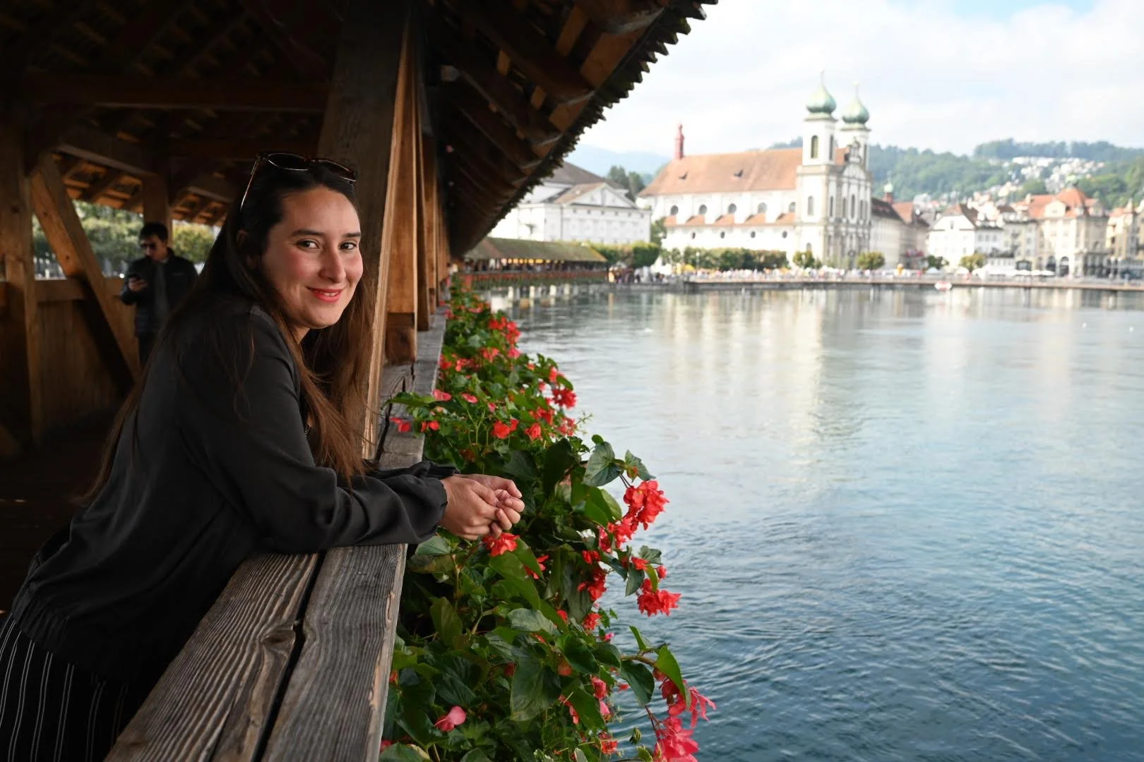 A woman with long brown hair smiling while leaning on a wooden railing decorated with pink flowers, with a body of water and historic buildings in the background.