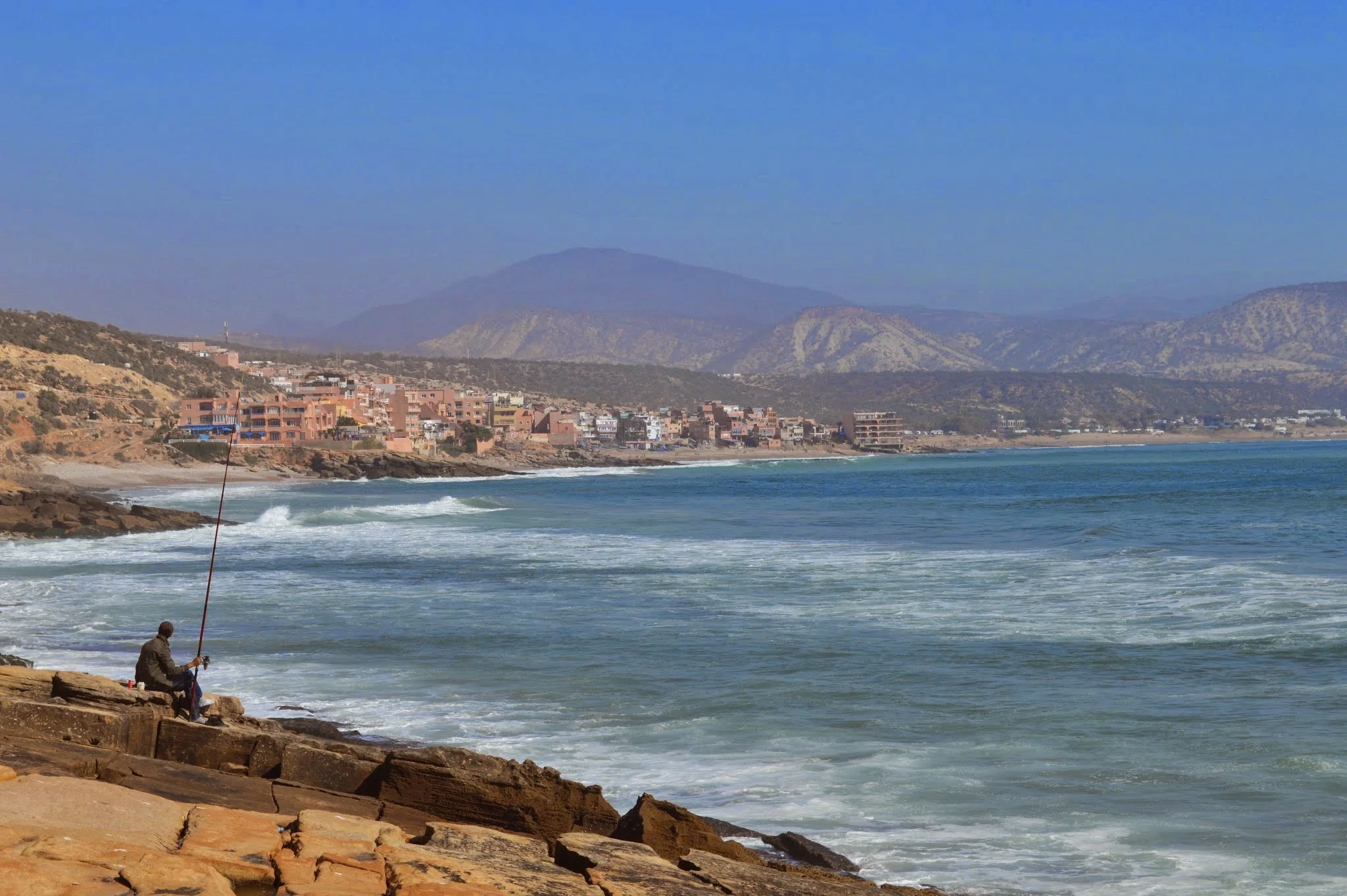 A man is sitting on rocks at the shoreline, fishing in the ocean with a city and mountains in the background.