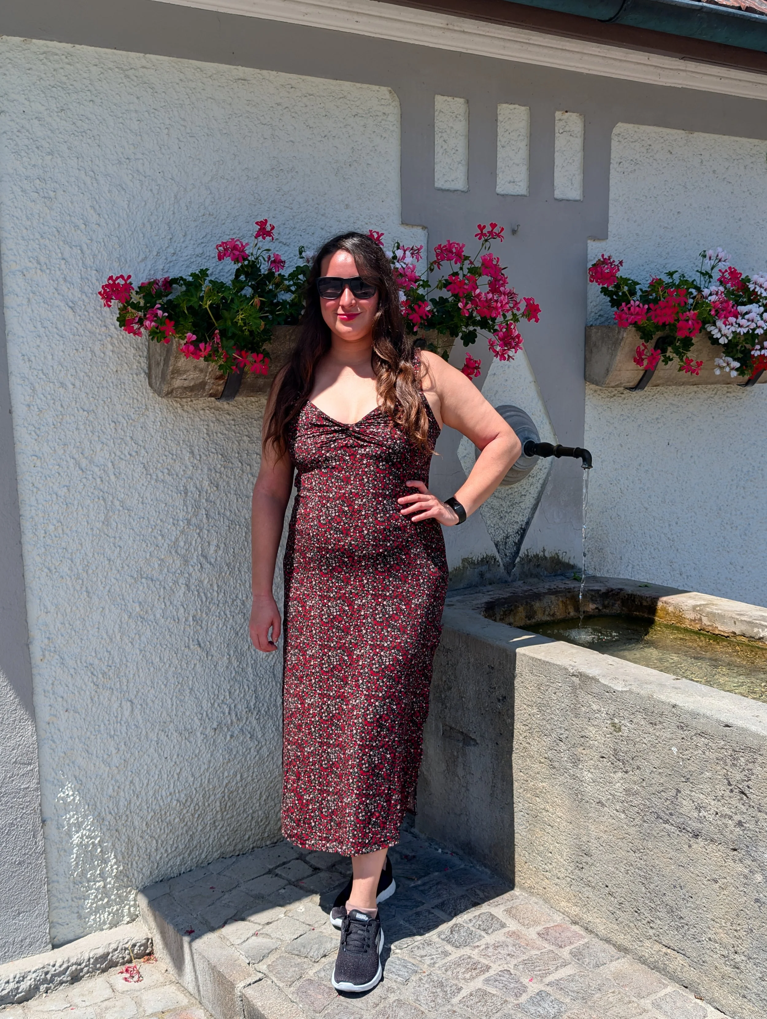 A woman with long dark hair, wearing sunglasses, a floral dress, and black sneakers, is standing in front of a white wall with pink and white flower planters and a water fountain.
