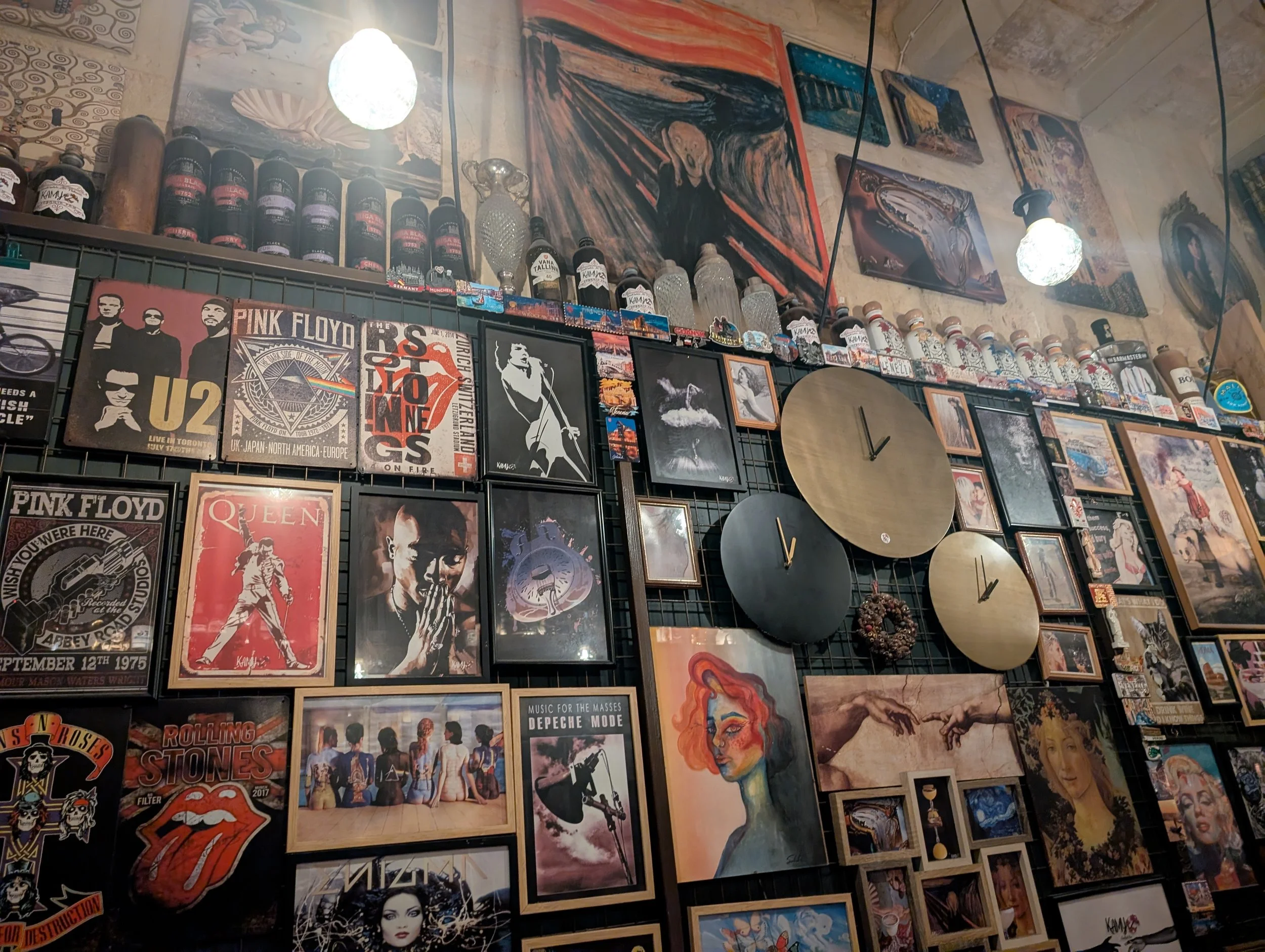 A wall decorated with framed posters, paintings, and other artwork, including images of famous bands such as Pink Floyd, Queen, and The Rolling Stones. There are also sculptures, mirrors, and hanging light fixtures.