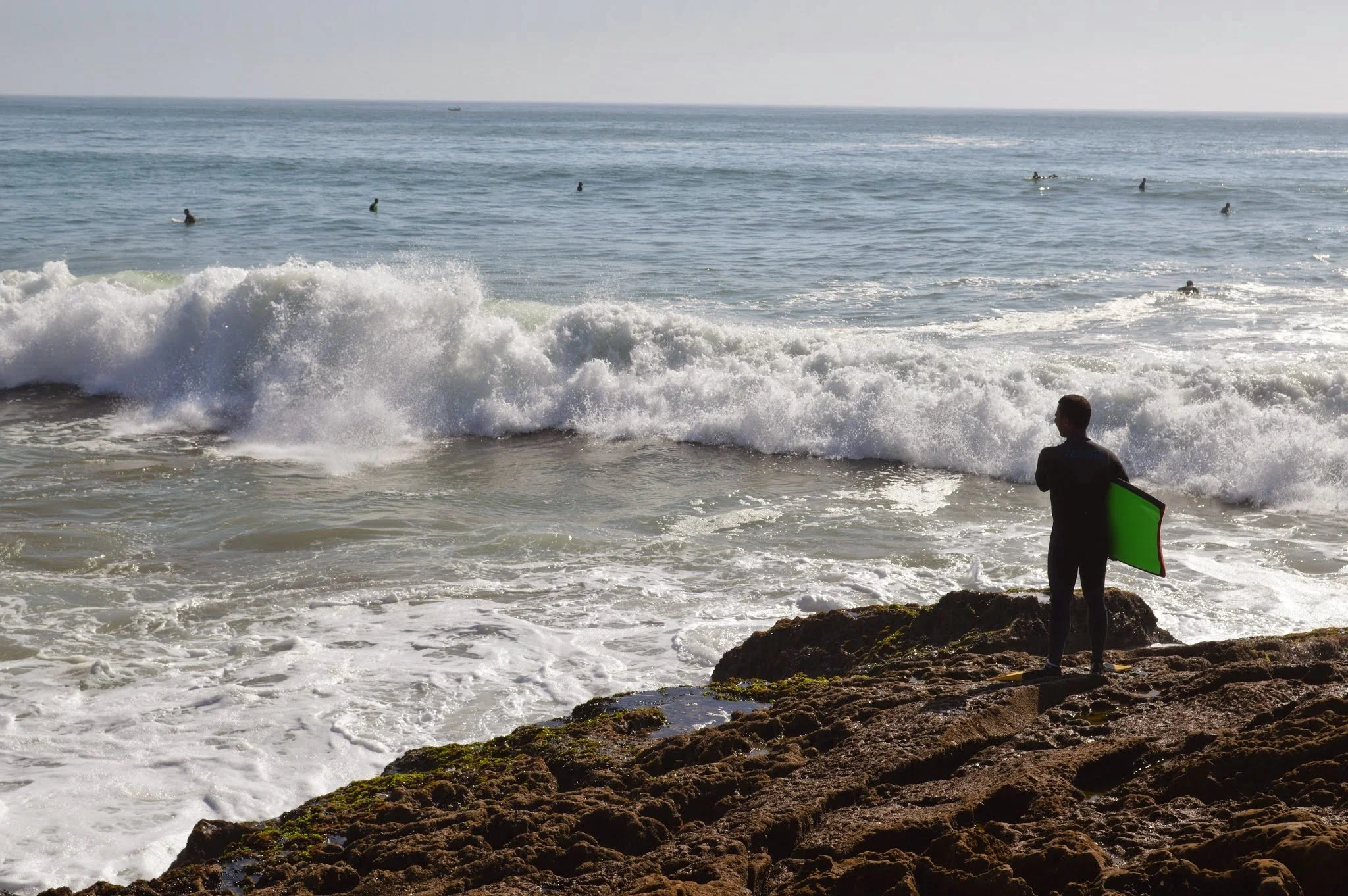 A person standing on rocky shore holding a green surfboard, watching surfers in the ocean waves.