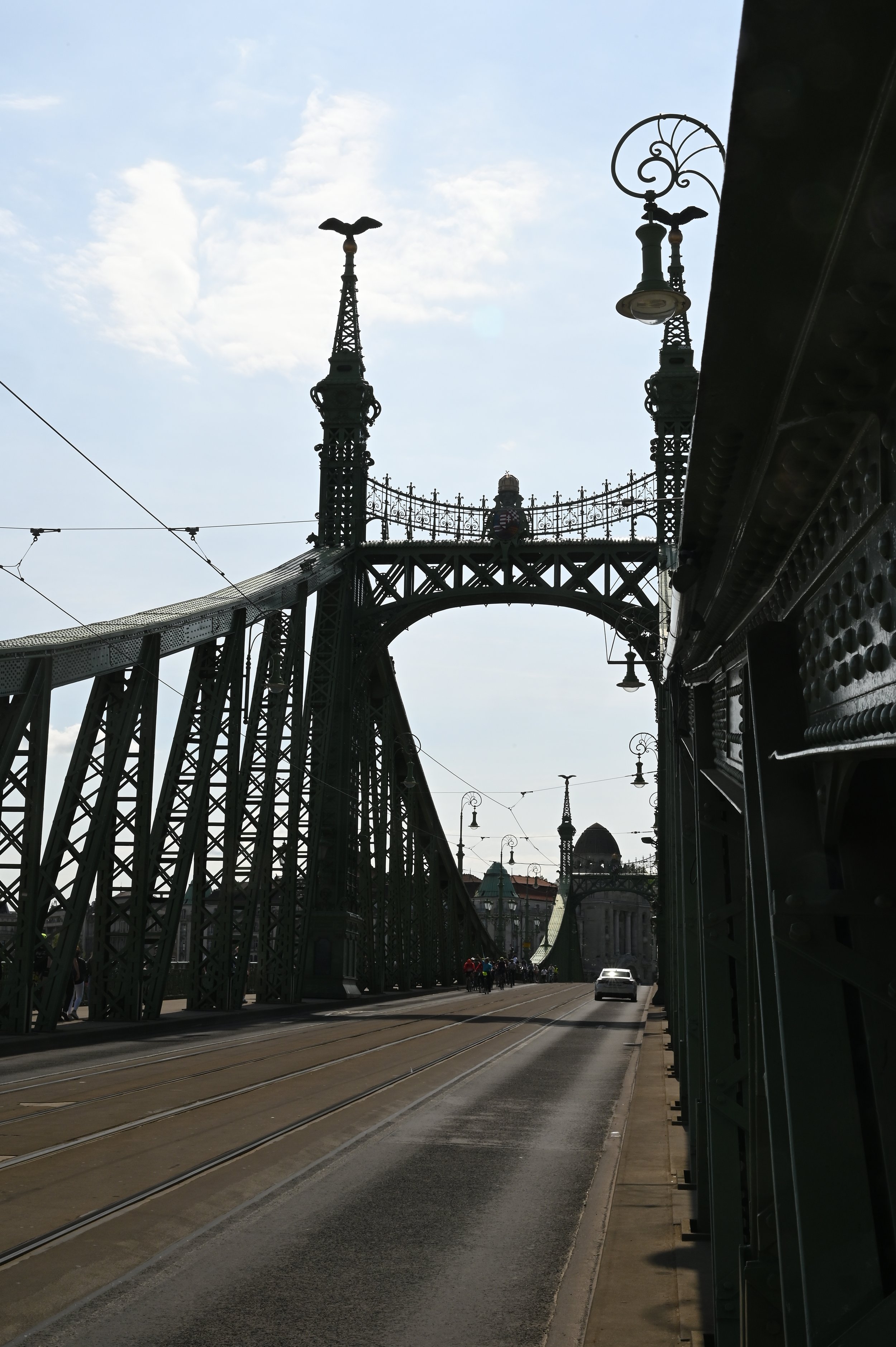 View of a bridge with decorative towers and street lights, tram tracks, and a few cars and pedestrians. The sky is partly cloudy.