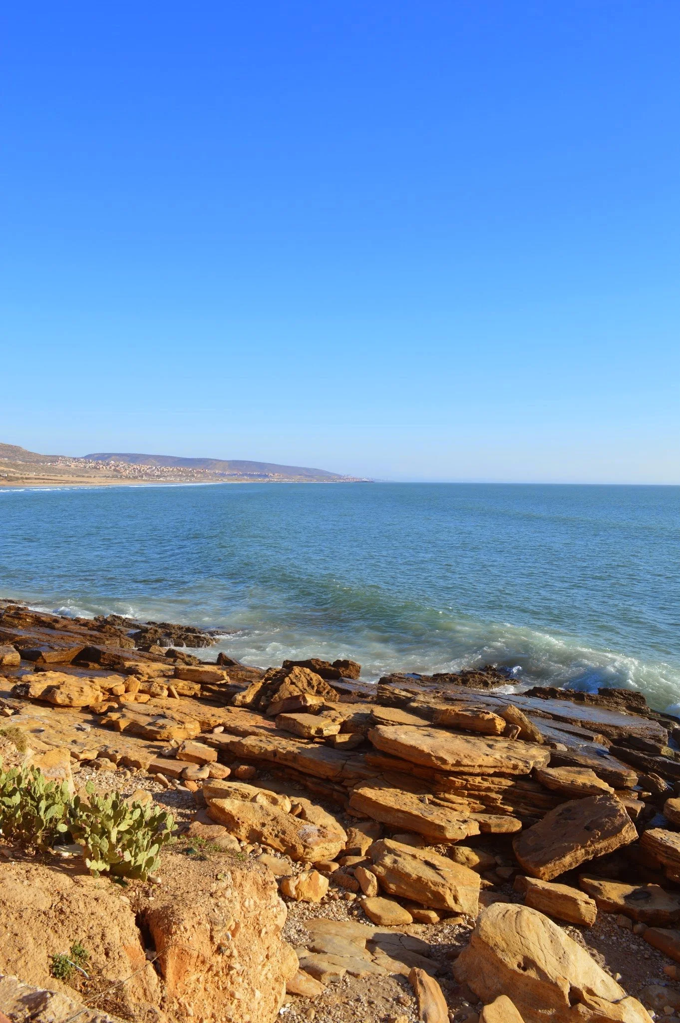 A rocky beach shoreline with large stones in the foreground, calm ocean waters in the middle ground, and distant hills with sparse buildings against a clear blue sky.