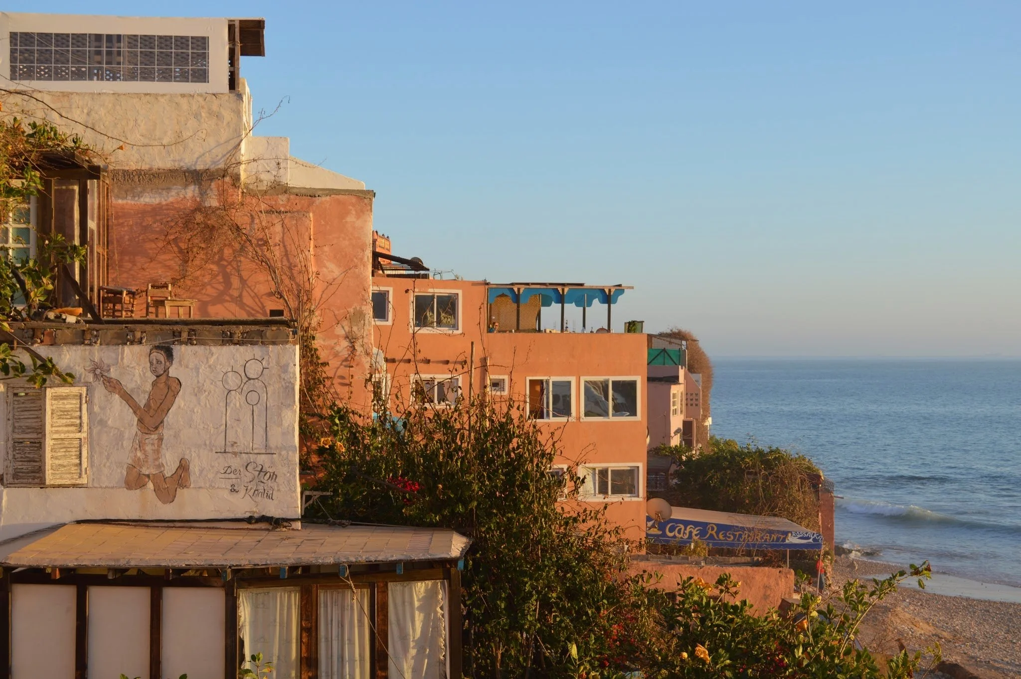 Colourful buildings on a coastal hillside with the ocean in the background, some buildings have balconies and veranda styles, and one has a mural of a woman holding flowers.