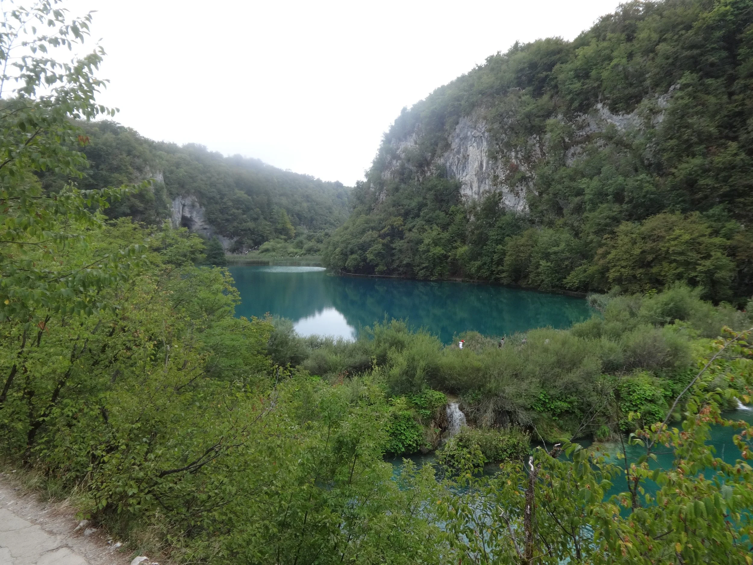 A scenic view of a turquoise river flowing through a lush green canyon with rocky cliffs on either side and trees overhanging the water, with a narrow dirt path at the lower left corner.