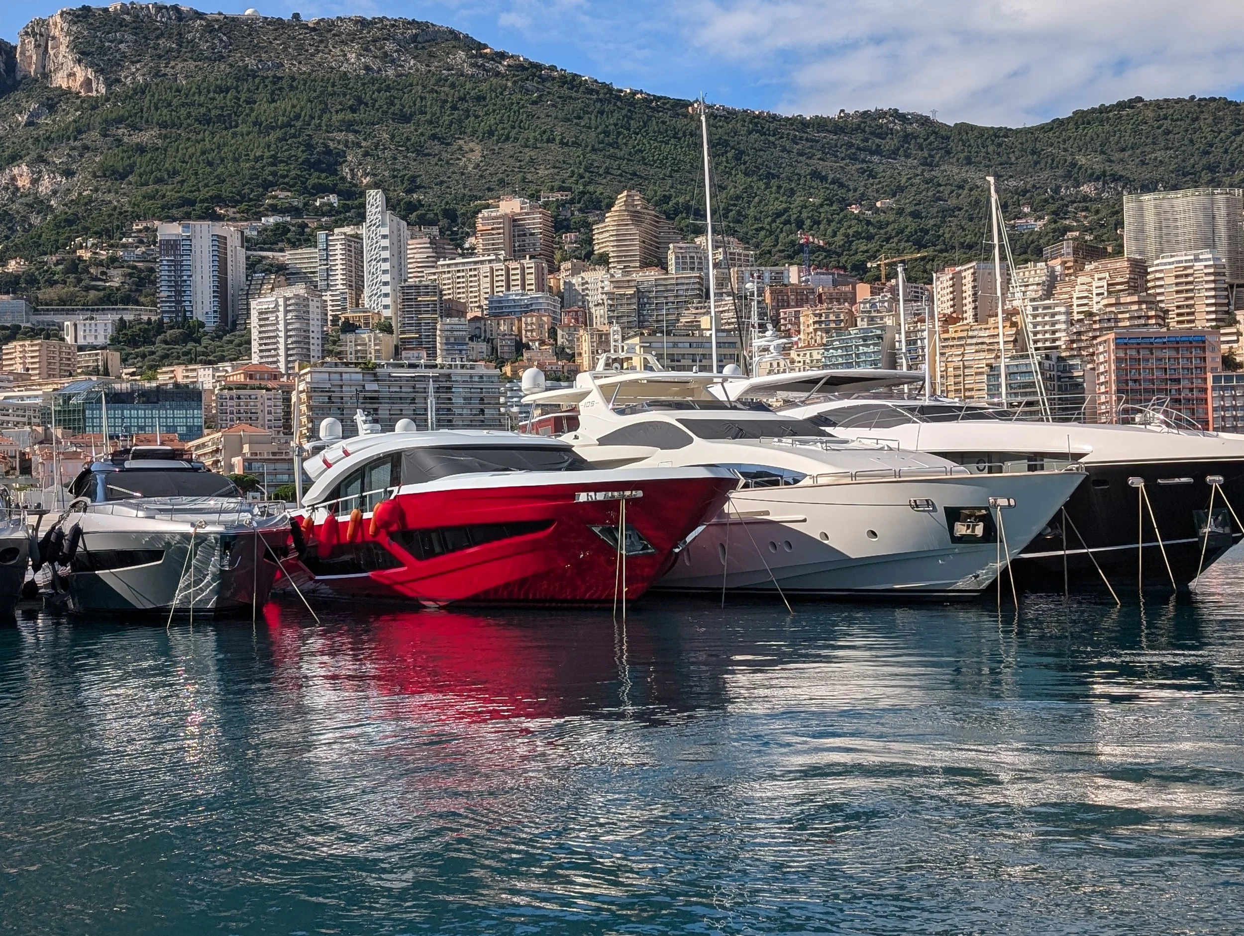 A marina with luxury yachts docked in the water, with a cityscape of high-rise buildings on a hillside and lush green mountains in the background.