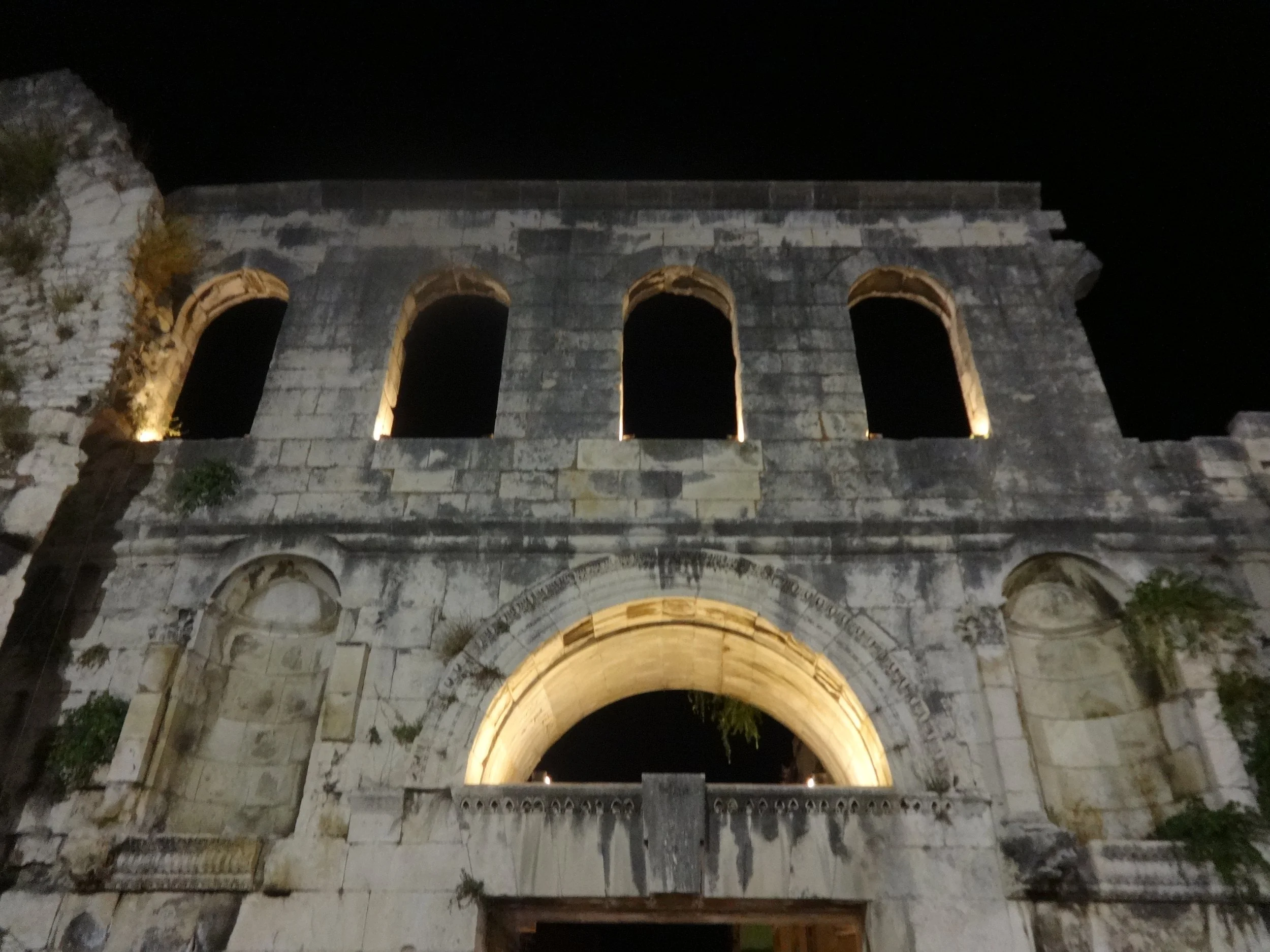 Night view of an ancient stone archway with three levels, illuminated from below, against a dark sky.