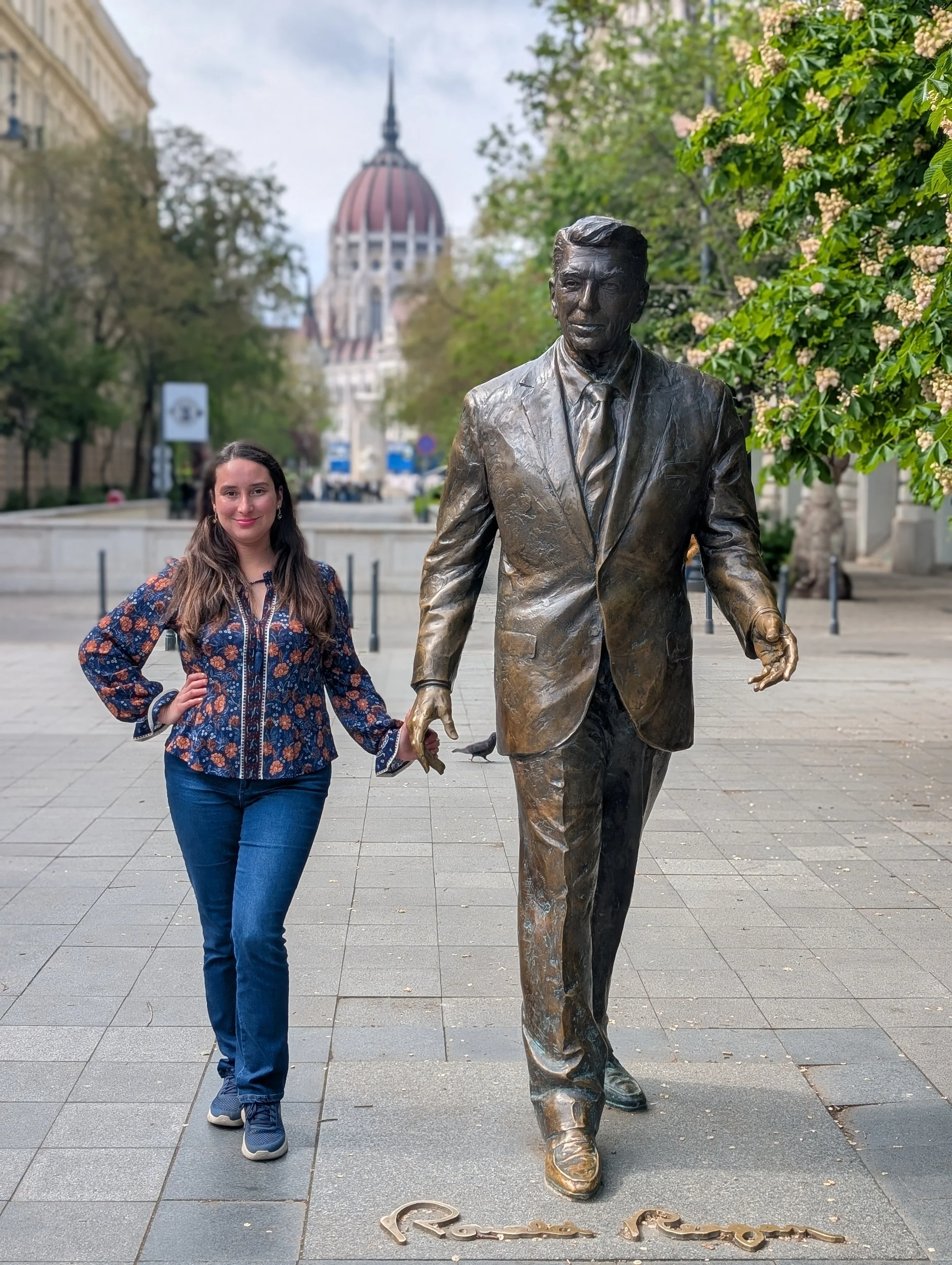 A woman standing next to a bronze statue of a man in a suit, with a historic building with a dome in the background and greenery around.