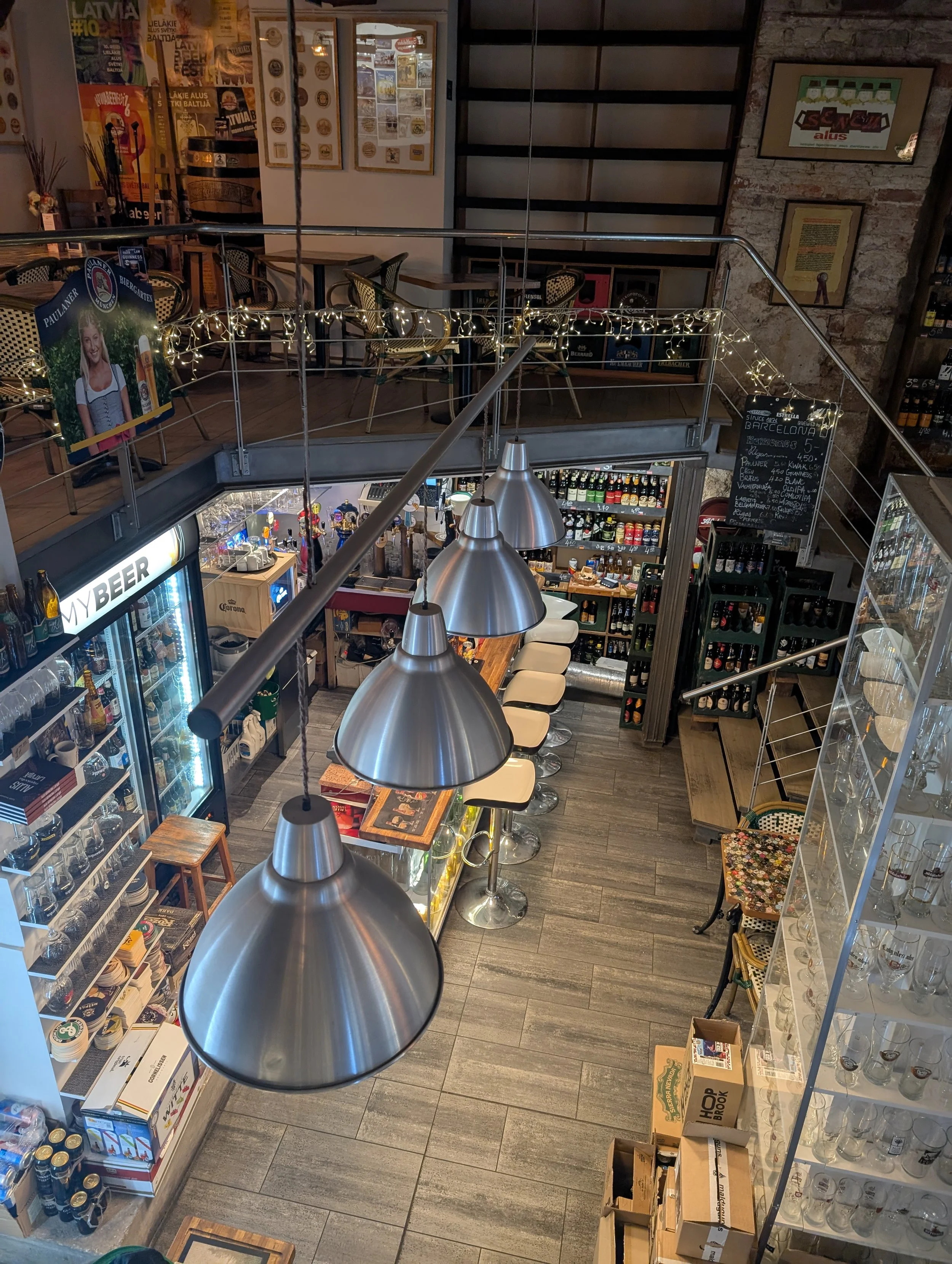 Interior of a bar or pub with shelves of bottles, a chalkboard menu, tables, chairs, and hanging lights.