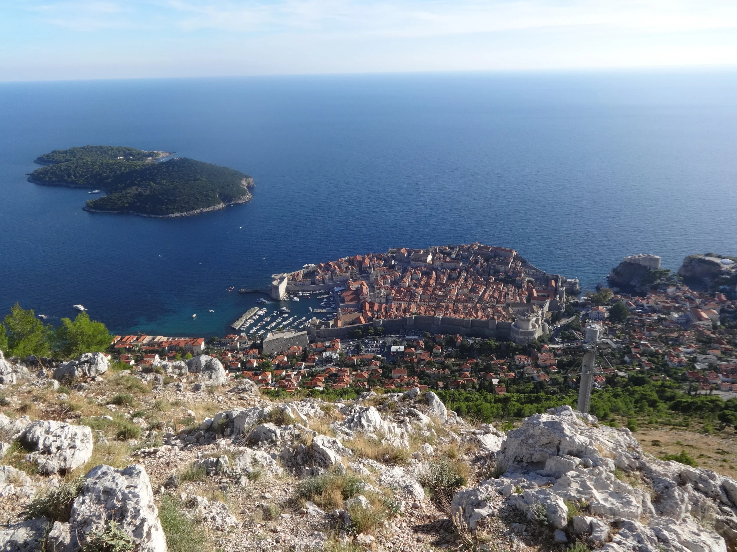 Aerial view of a coastal town with red rooftops, surrounded by a harbor with boats, and an island covered in greenery in the sea. Rocky terrain with sparse vegetation in the foreground.