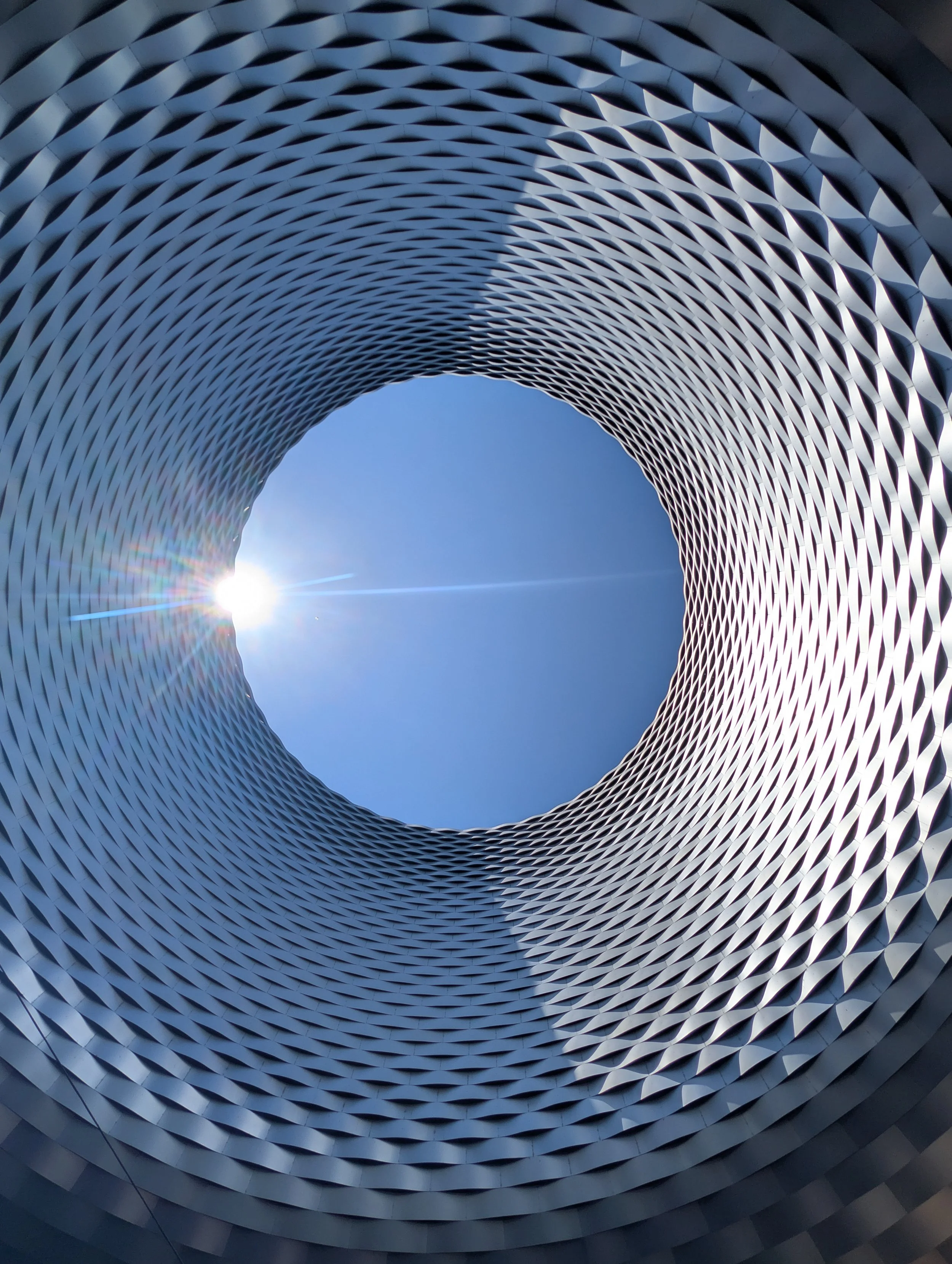 Looking up through the circular patterned windows of a modern architectural building towards a clear blue sky with the sun visible at the bottom of the frame.