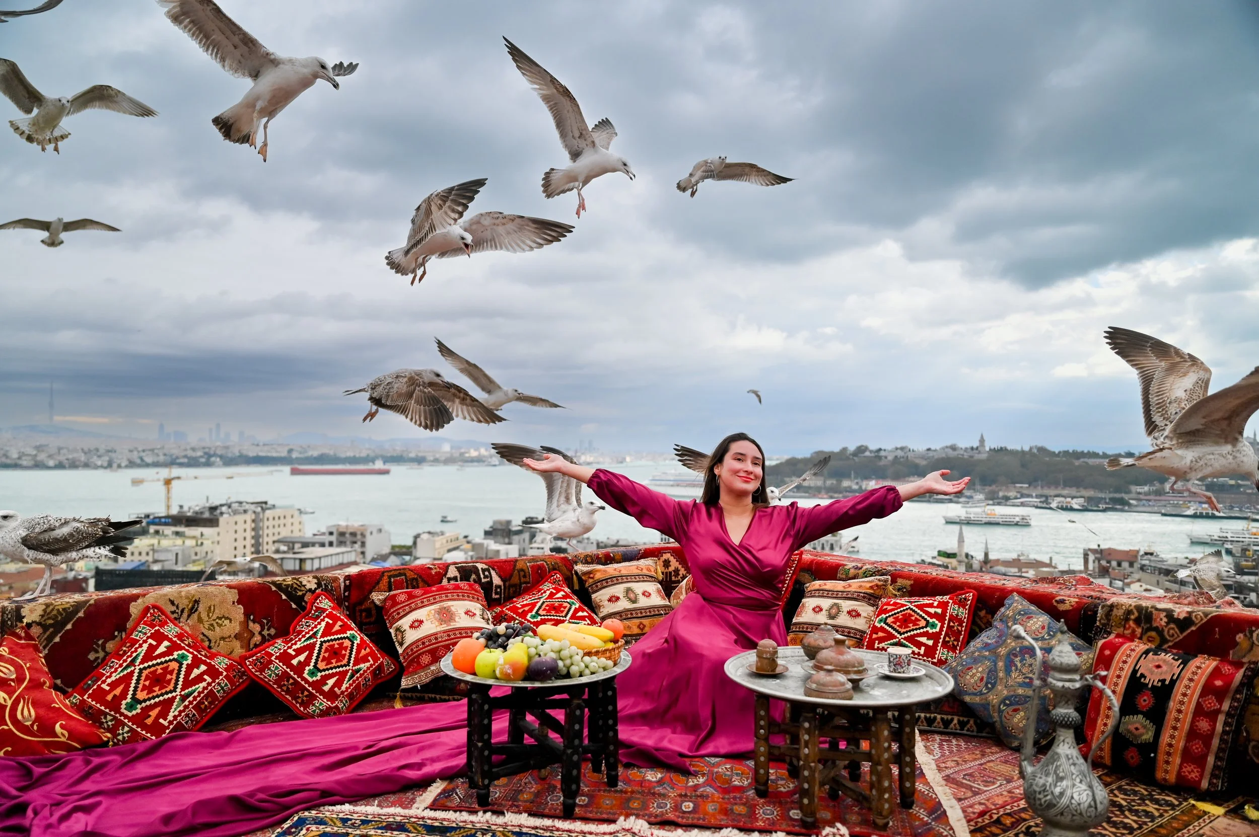 A woman in a pink traditional dress is sitting on a colorful, patterned carpet on a rooftop with a city port in the background. She is smiling with her arms outstretched as seagulls fly around her. There are plates of fruits and traditional items on 
