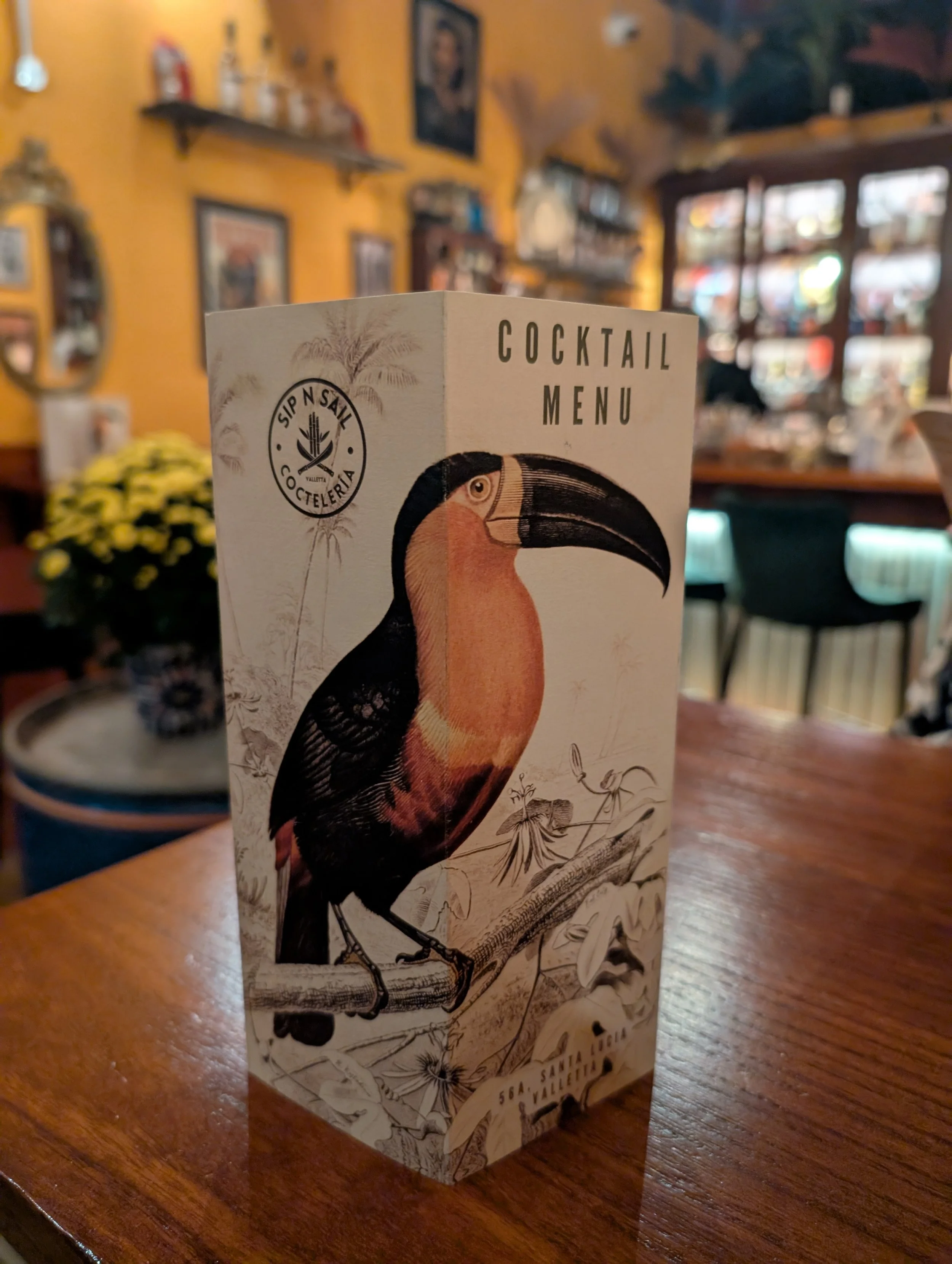 A cocktail menu with a toucan illustration inside a restaurant with warm lighting, wooden tables, and decorated walls.