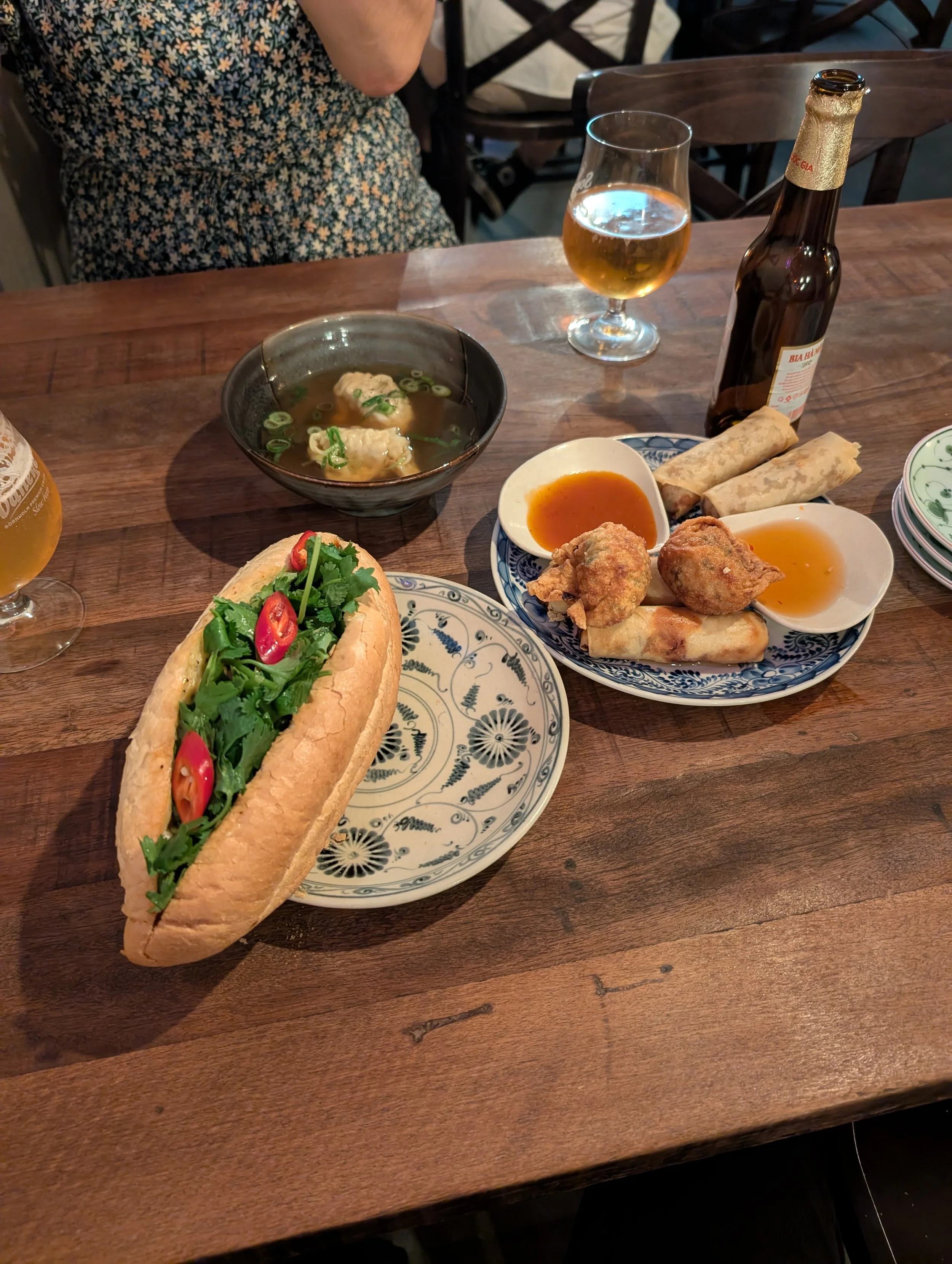 A wooden table with various Asian dishes, including a large banh mi sandwich with vegetables, a bowl of wonton soup, spring rolls, fried food with dipping sauces, a glass of beer, and an empty bottle of beer, in a restaurant setting.