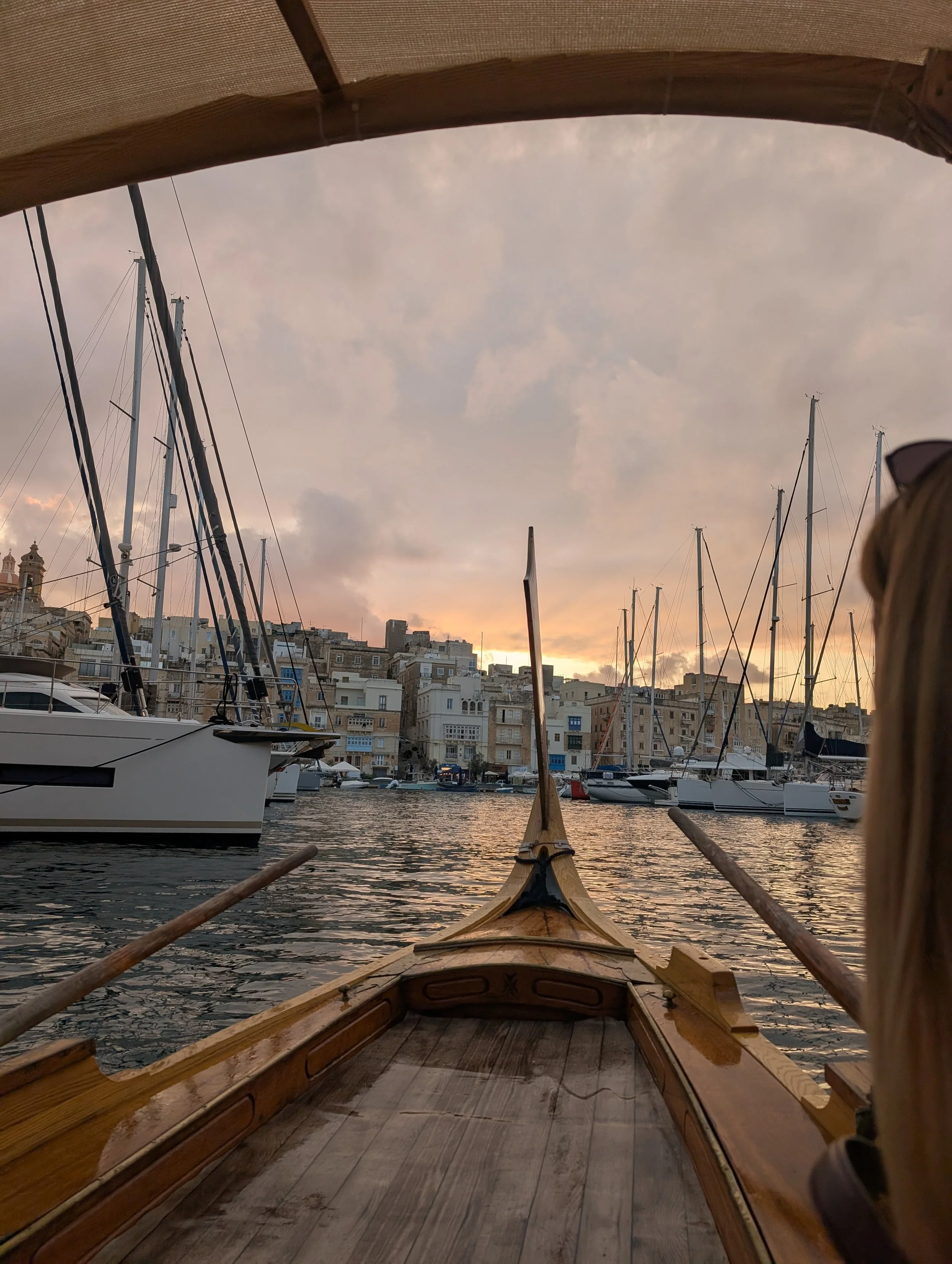 View from a small wooden boat on a harbor at sunset, with numerous sailboats and yachts docked and a cityscape in the background under a cloudy sky.
