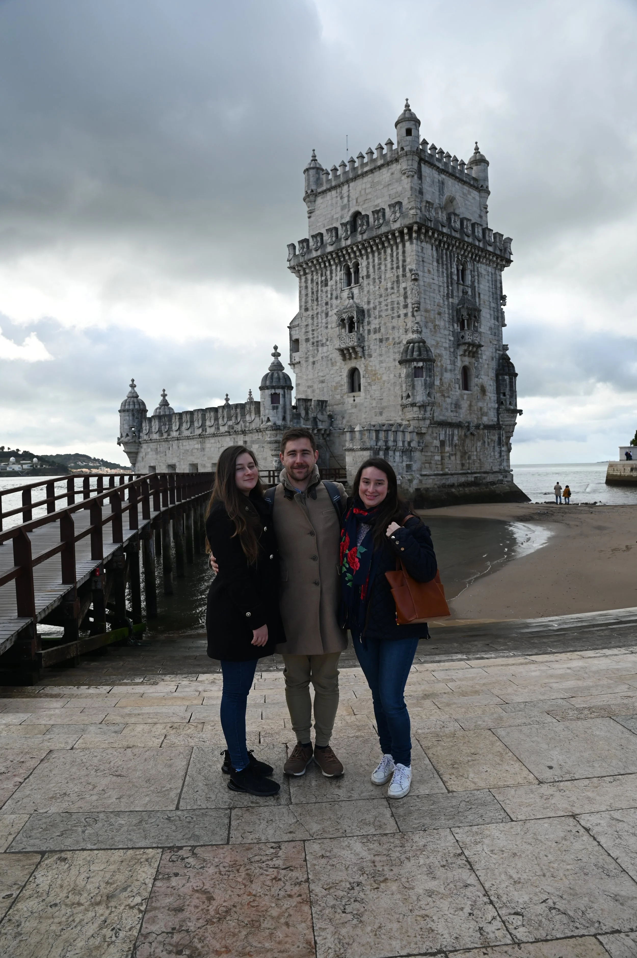Three people standing on a stone-paved walkway in front of the Tower of Belém, a historic fortress with a white stone facade, small towers, and battlements, overlooking the river, under a cloudy sky.