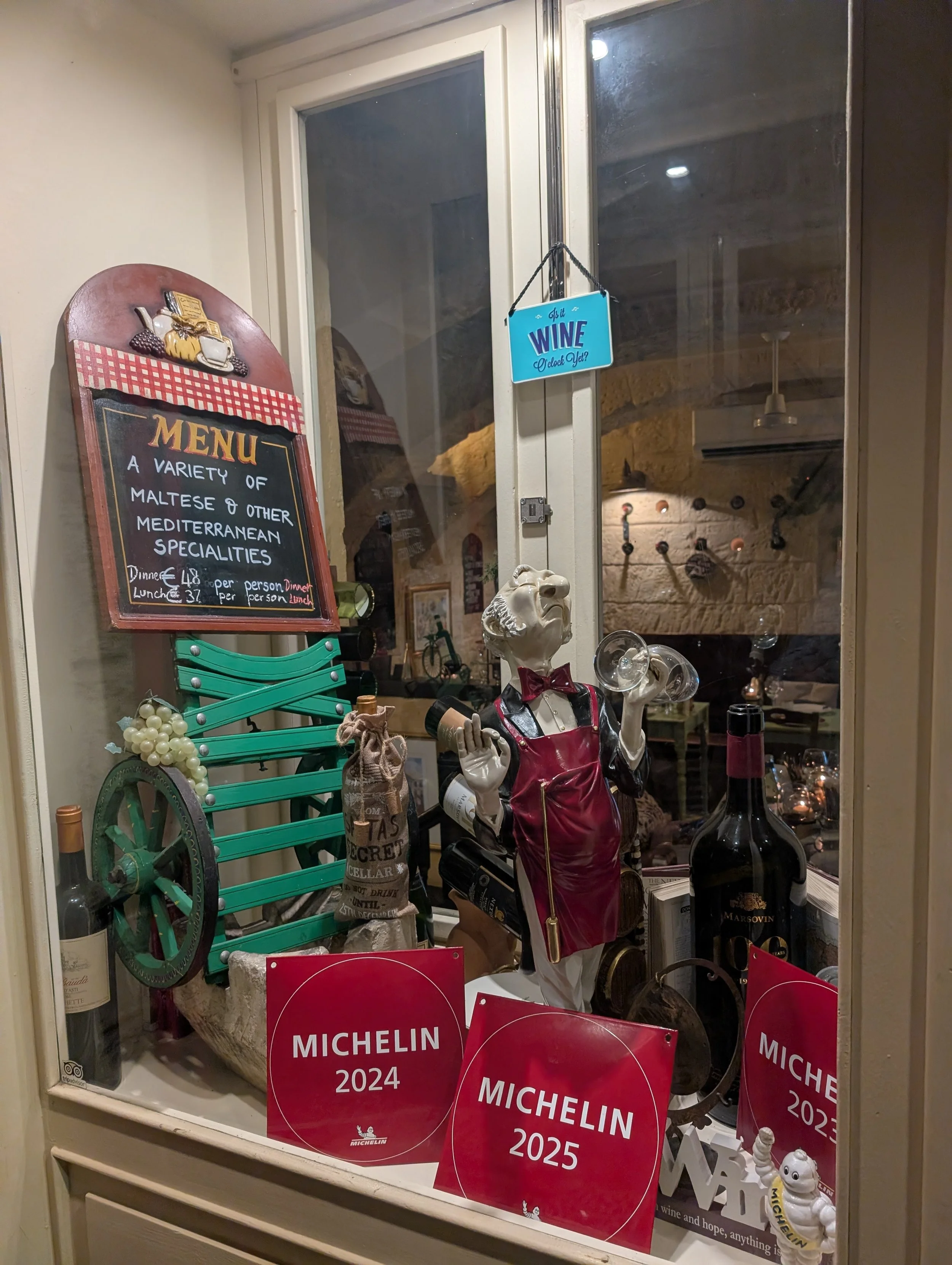 View of a restaurant or cafe window display with a colorful menu board, a decorative wine-themed sign hanging outside, and various decorative items including a skeleton figure dressed as a waiter, wine bottles, and red Michelin awards for 2024, 2025,