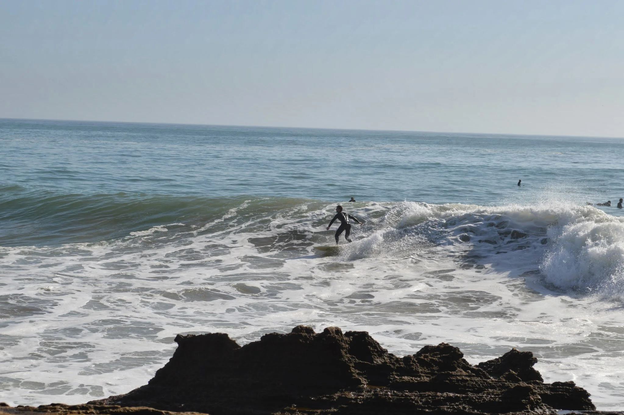 A person surfing on a wave in the ocean, with rocks in the foreground and other surfers in the water, under a clear sky.