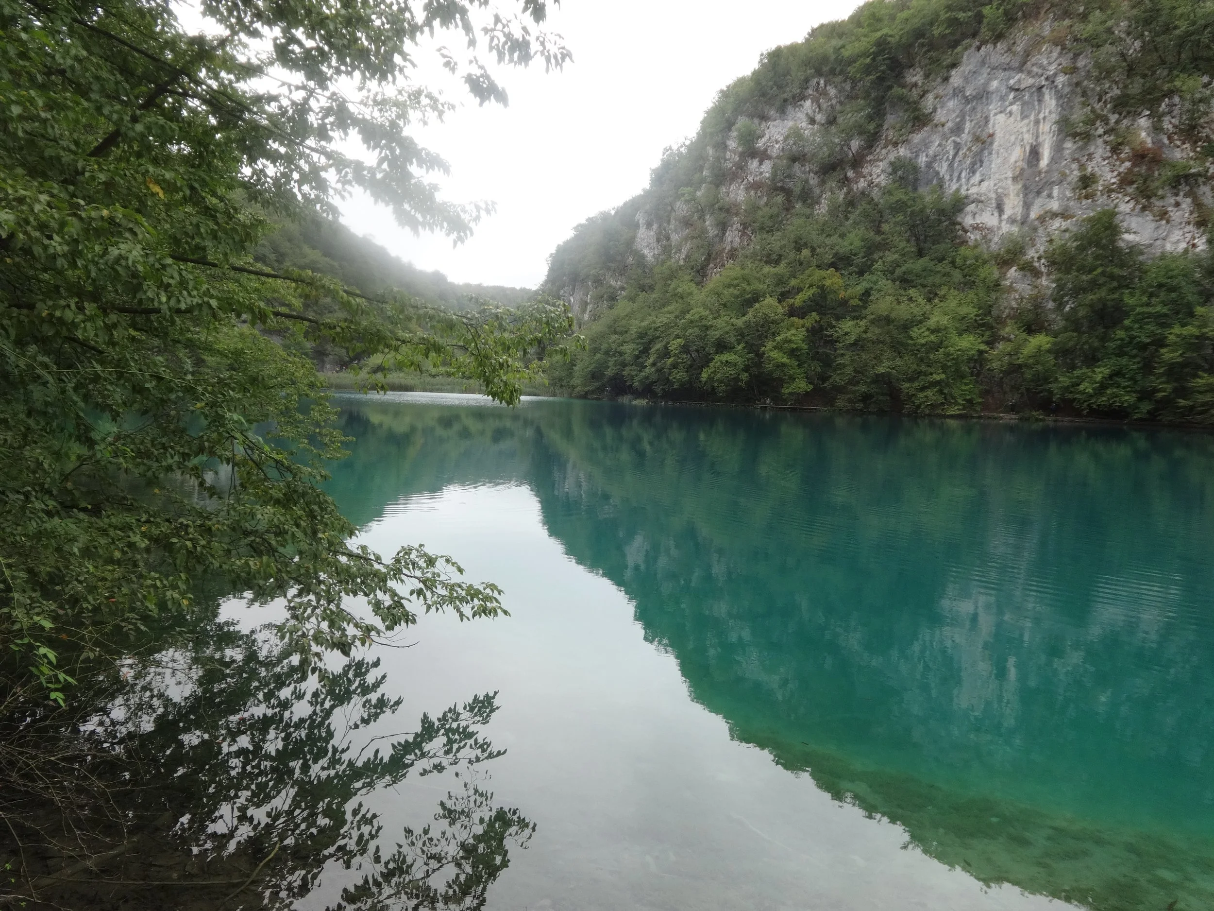 A serene landscape featuring a body of turquoise water surrounded by green trees and a steep rocky hillside under a misty sky.