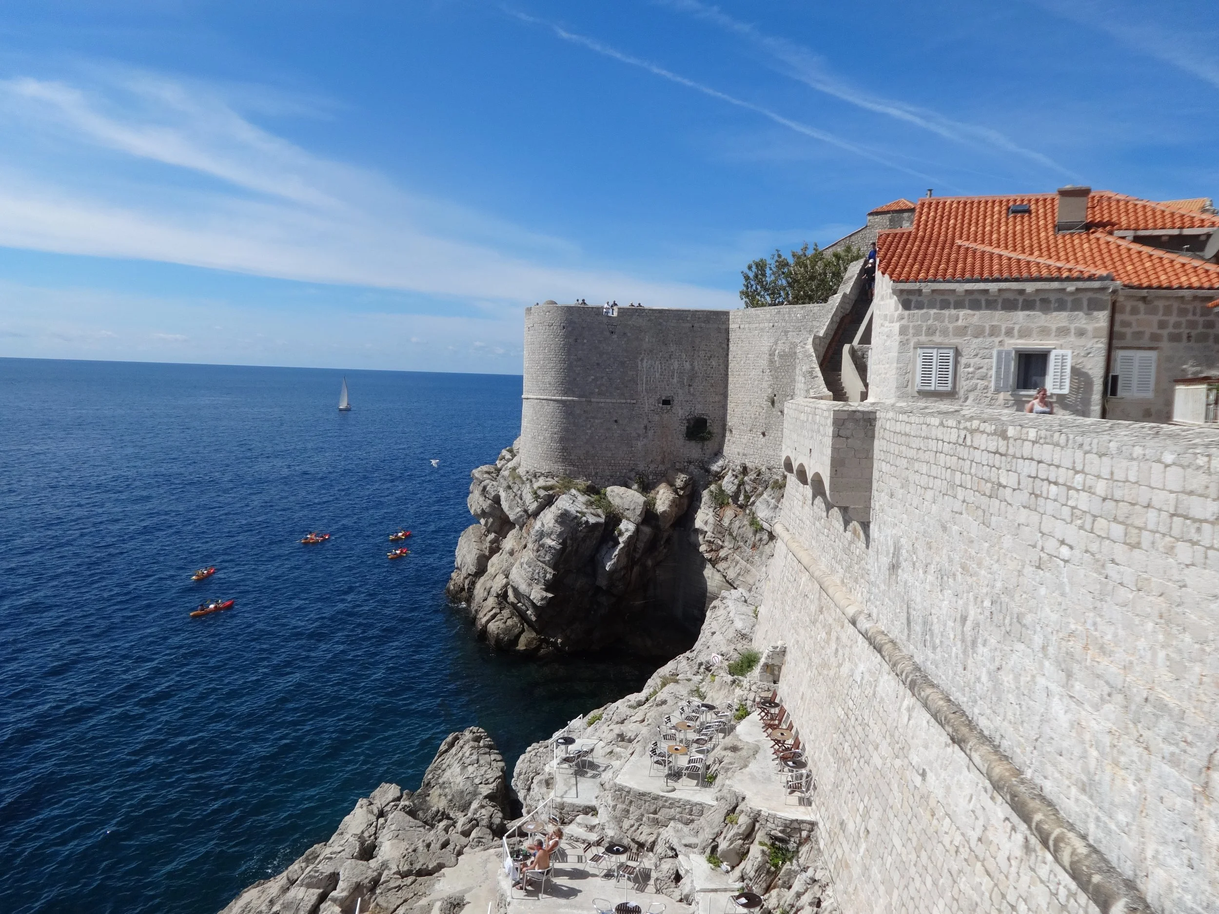A coastal scene with a stone fortress or castle wall extending into the sea under a blue sky with wispy clouds. There are people on the wall and some sitting at tables along a stone walkway, with several kayaks in the water and a sailboat on the hori