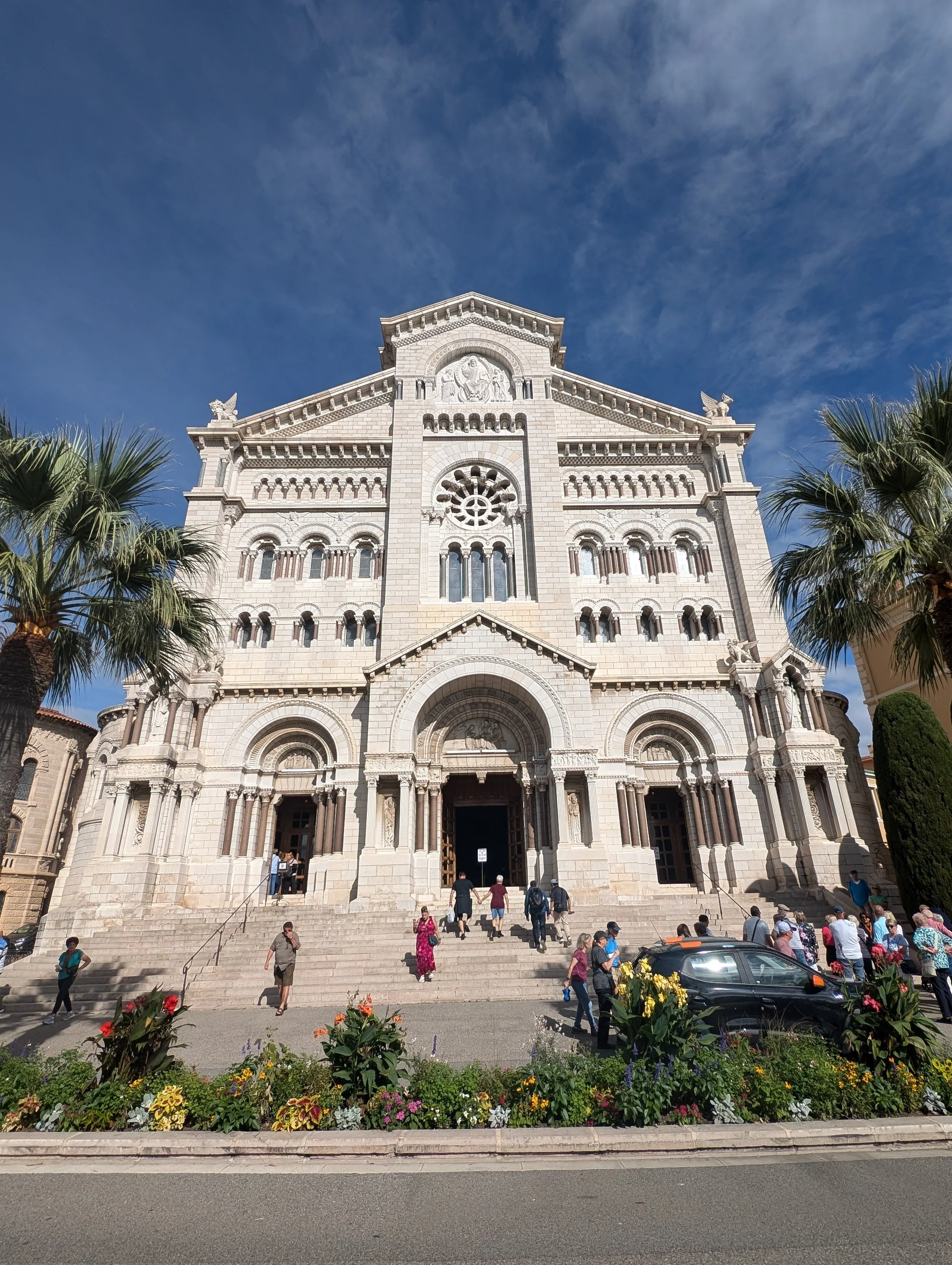People walking up and down the steps of a large, historic cathedral with intricate stone architecture, sculptures, and stained glass windows, surrounded by palm trees and flower beds.