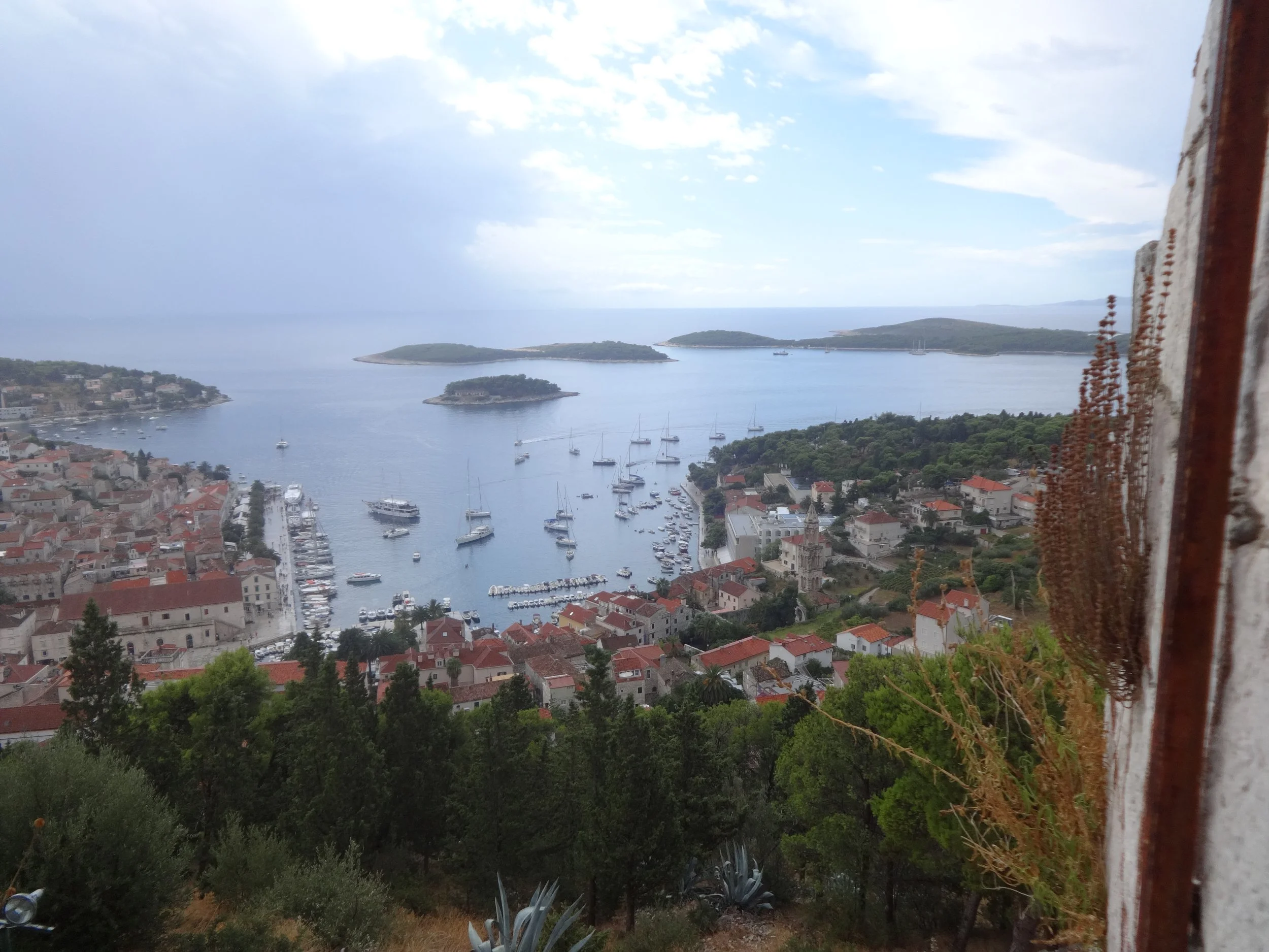 A view of a coastal town with red-roofed buildings, boats in the harbor, and small islands in the water, seen from a high vantage point.