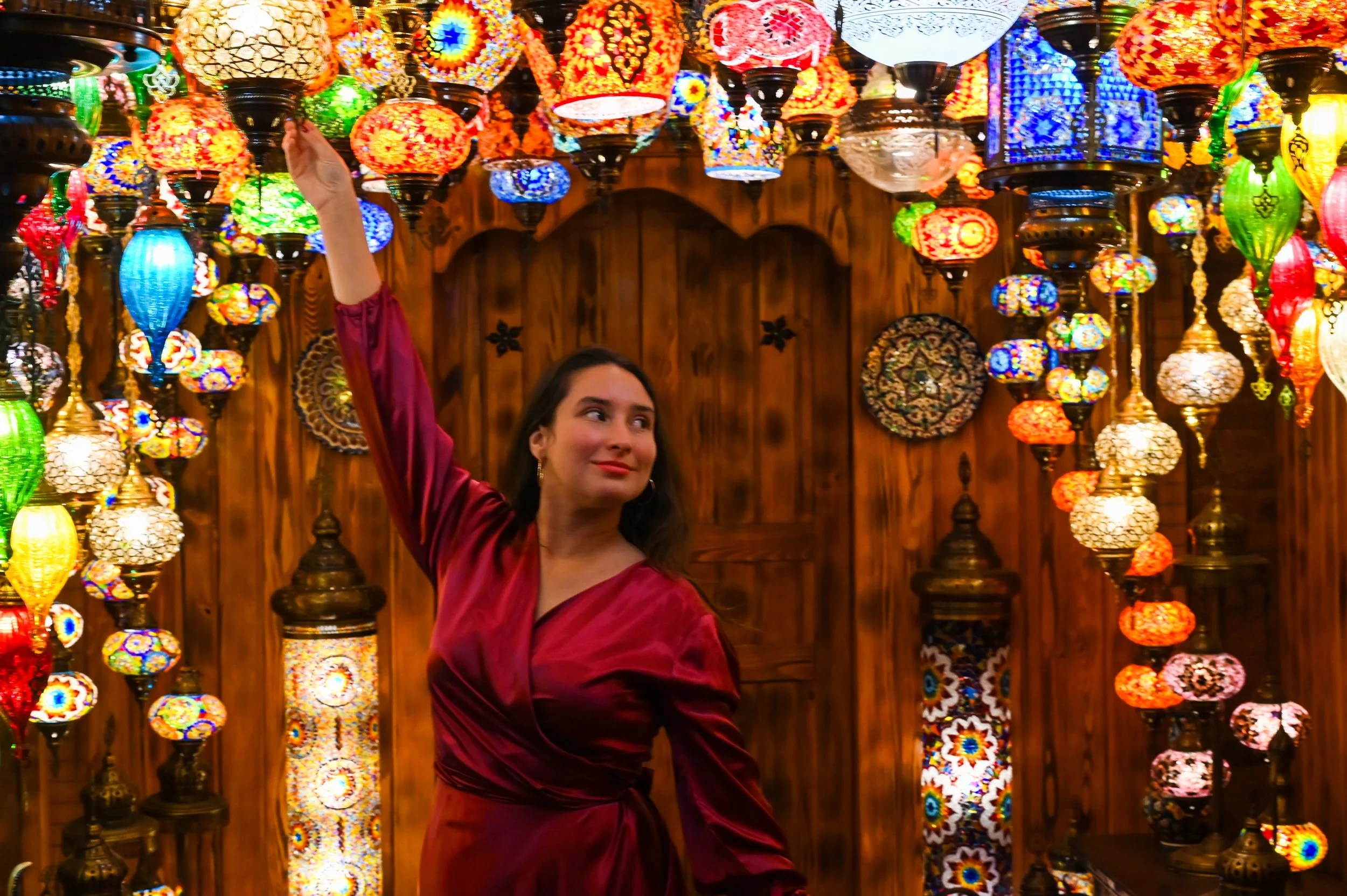 A woman in a red satin dress surrounded by colorful, ornate Turkish lamps hanging from the ceiling of a wooden interior.