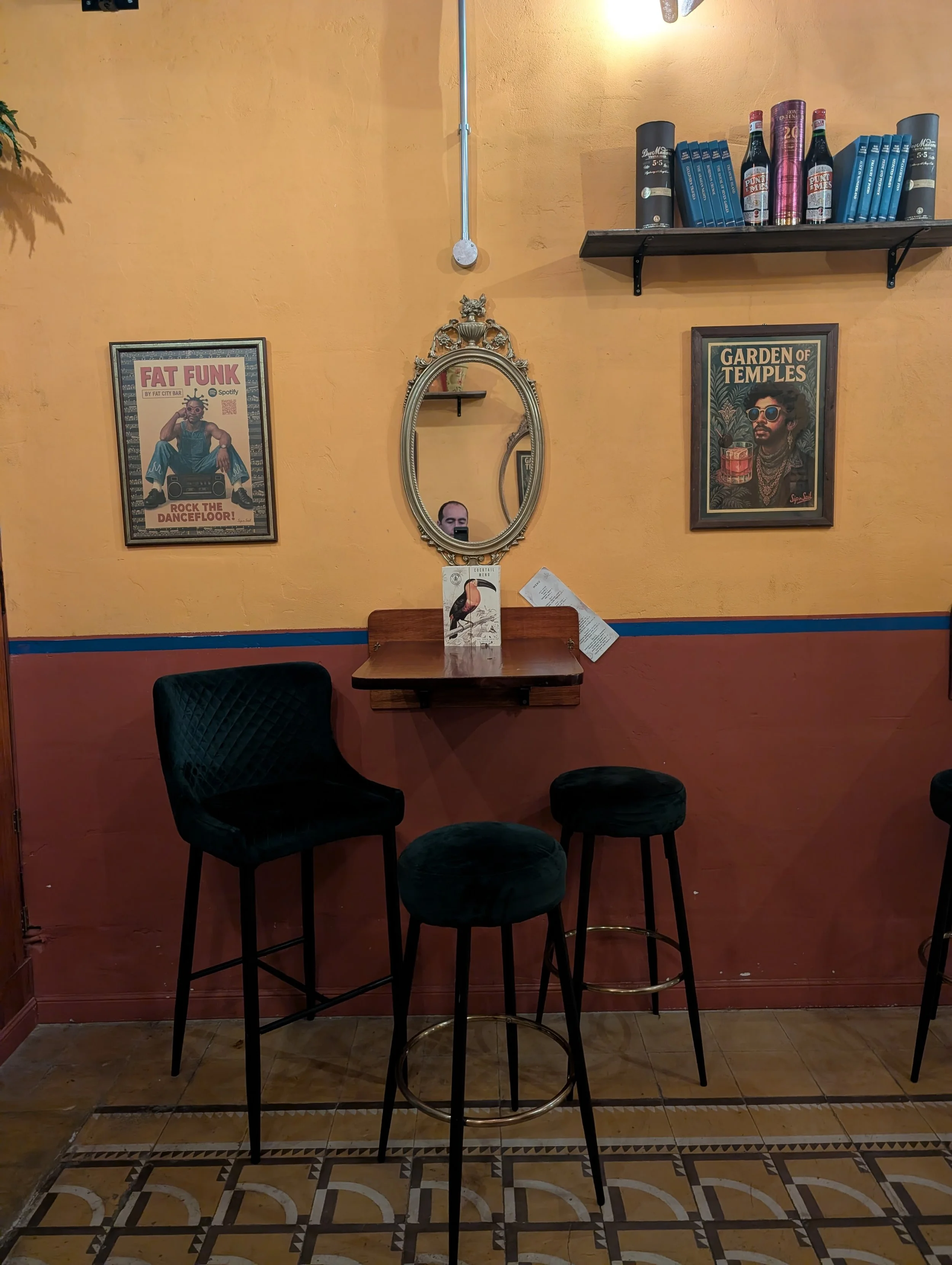 Image of a restaurant or cafe interior featuring a yellow and orange wall with decorative framed posters, a small ornate mirror, a small shelf with a menu, and tall black chairs with plush seats positioned around a wooden table. The floor has pattern