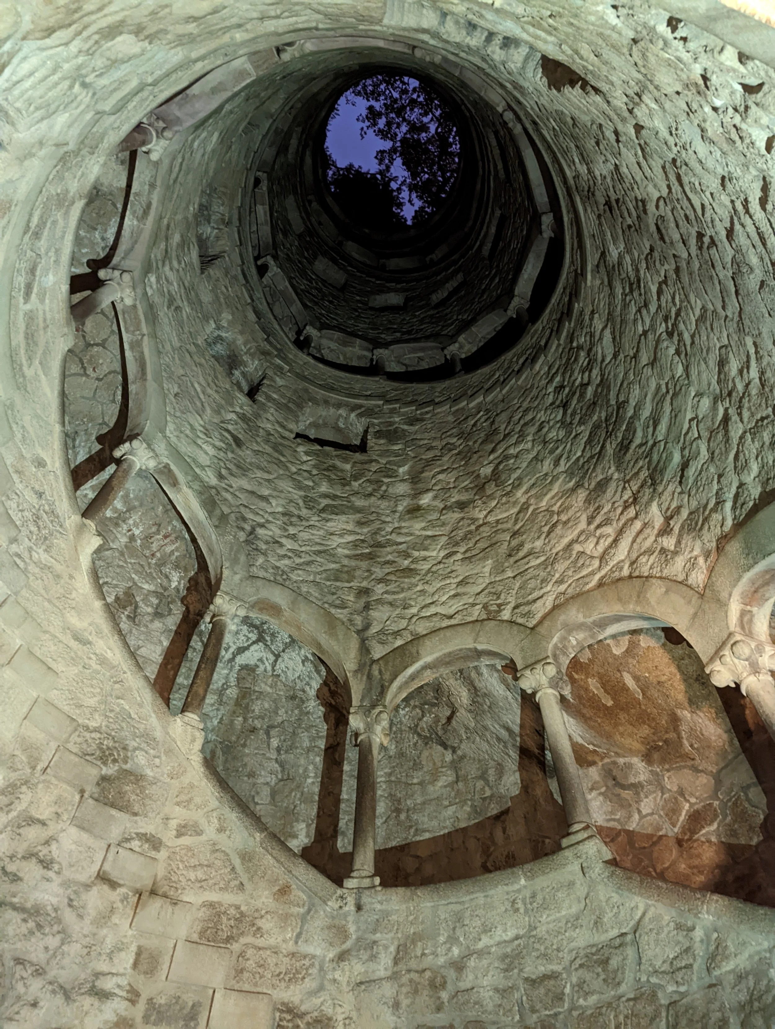 A view looking up inside a stone tower or well, showing a spiral staircase and stone walls, with a view of the sky and tree branches at the top.