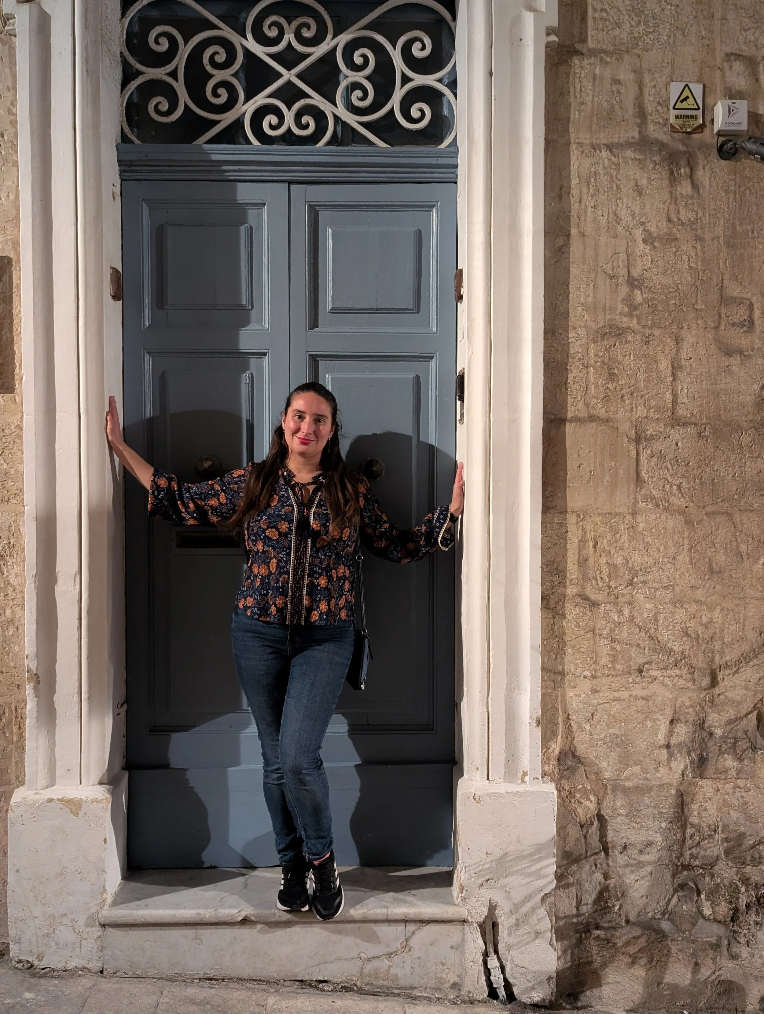 A woman standing in front of a large gray door with decorative ironwork above it. She is smiling, wearing a patterned blouse, jeans, and sneakers, and has her arms extended to hold onto the sides of the door frame.