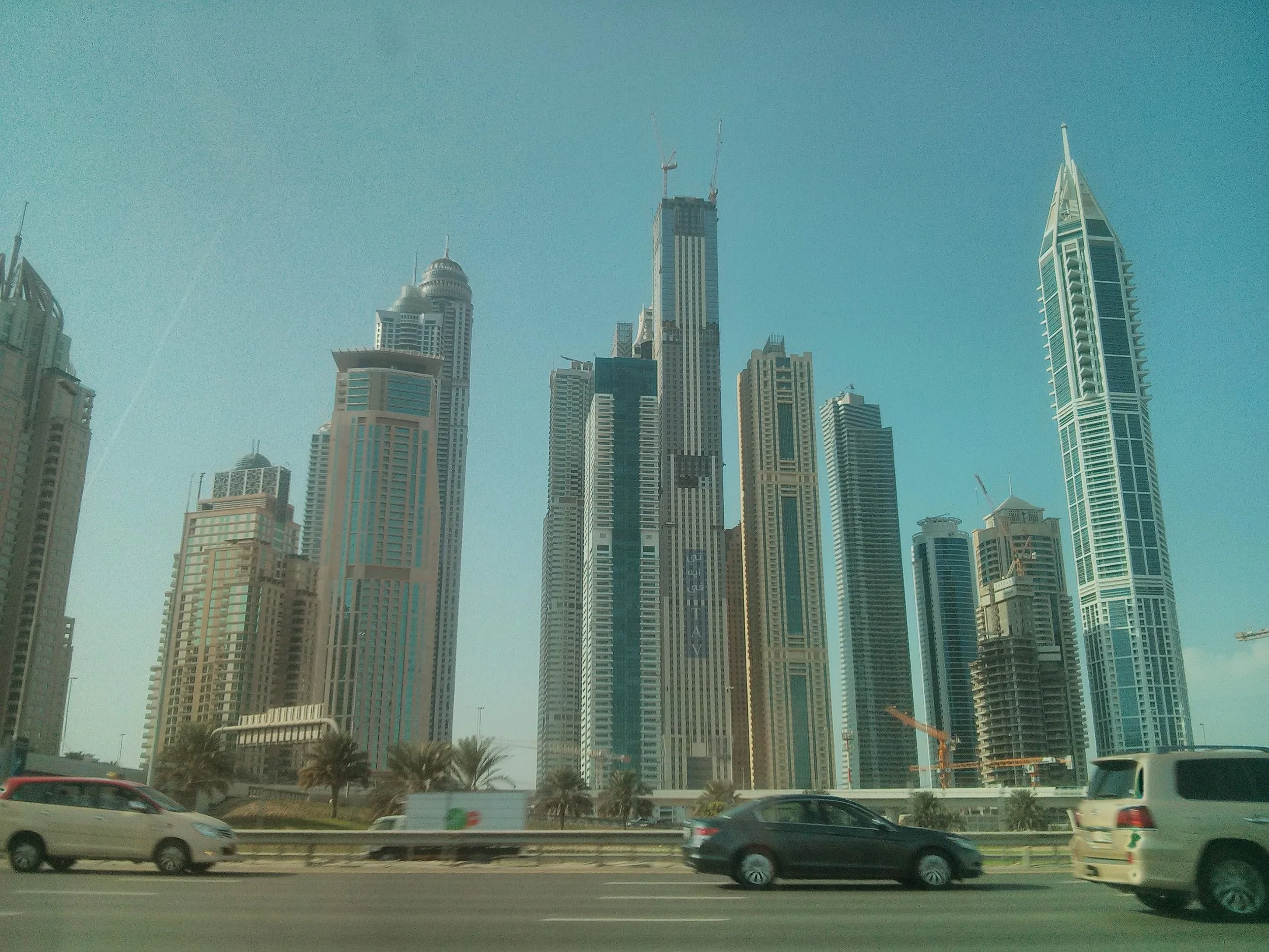 Skyline of tall modern skyscrapers in a city with clear blue sky, viewed from a highway with cars in the foreground.