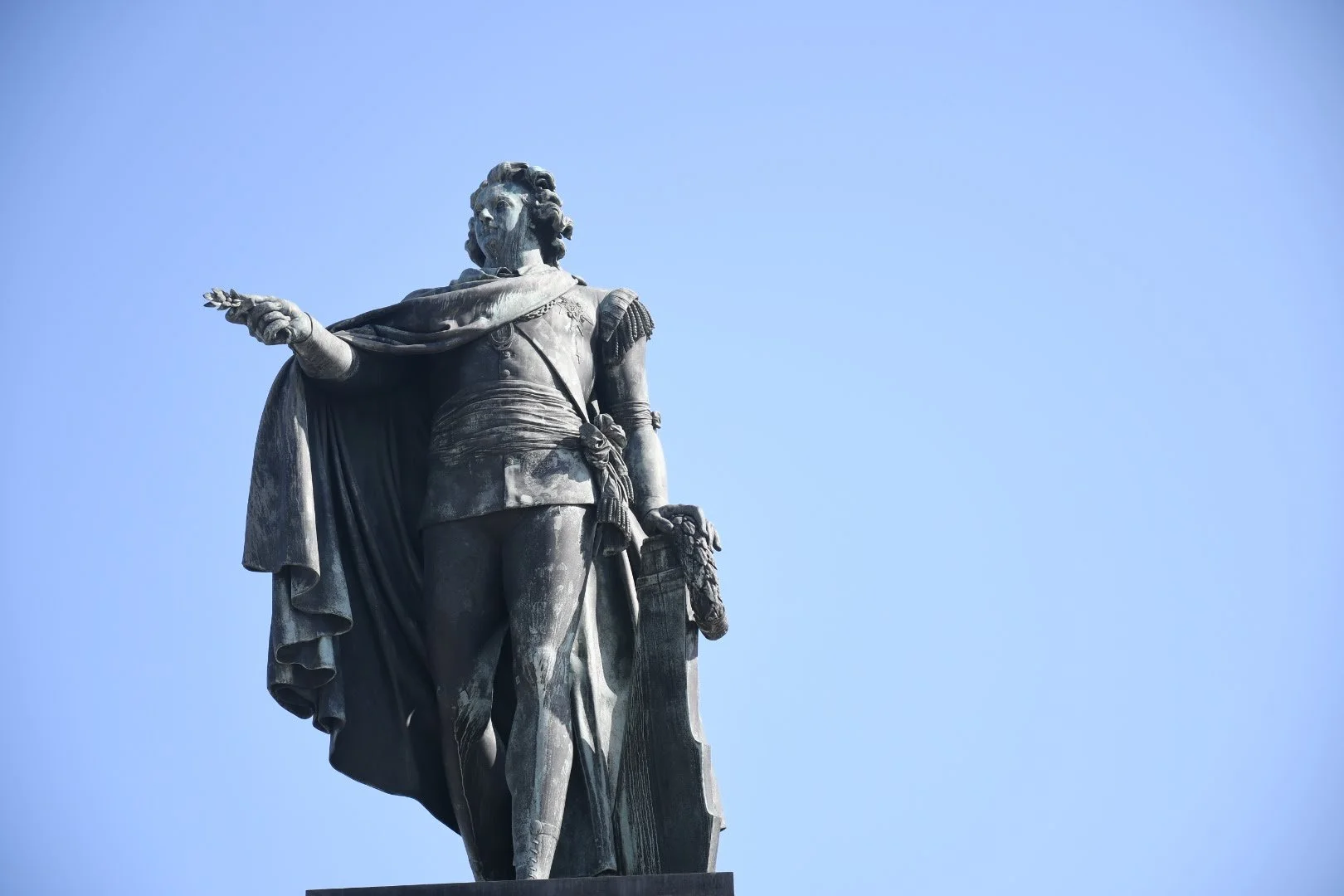 Statue of a historical figure in military attire, pointing forward, with a cape draped over the shoulder, set against a clear blue sky.
