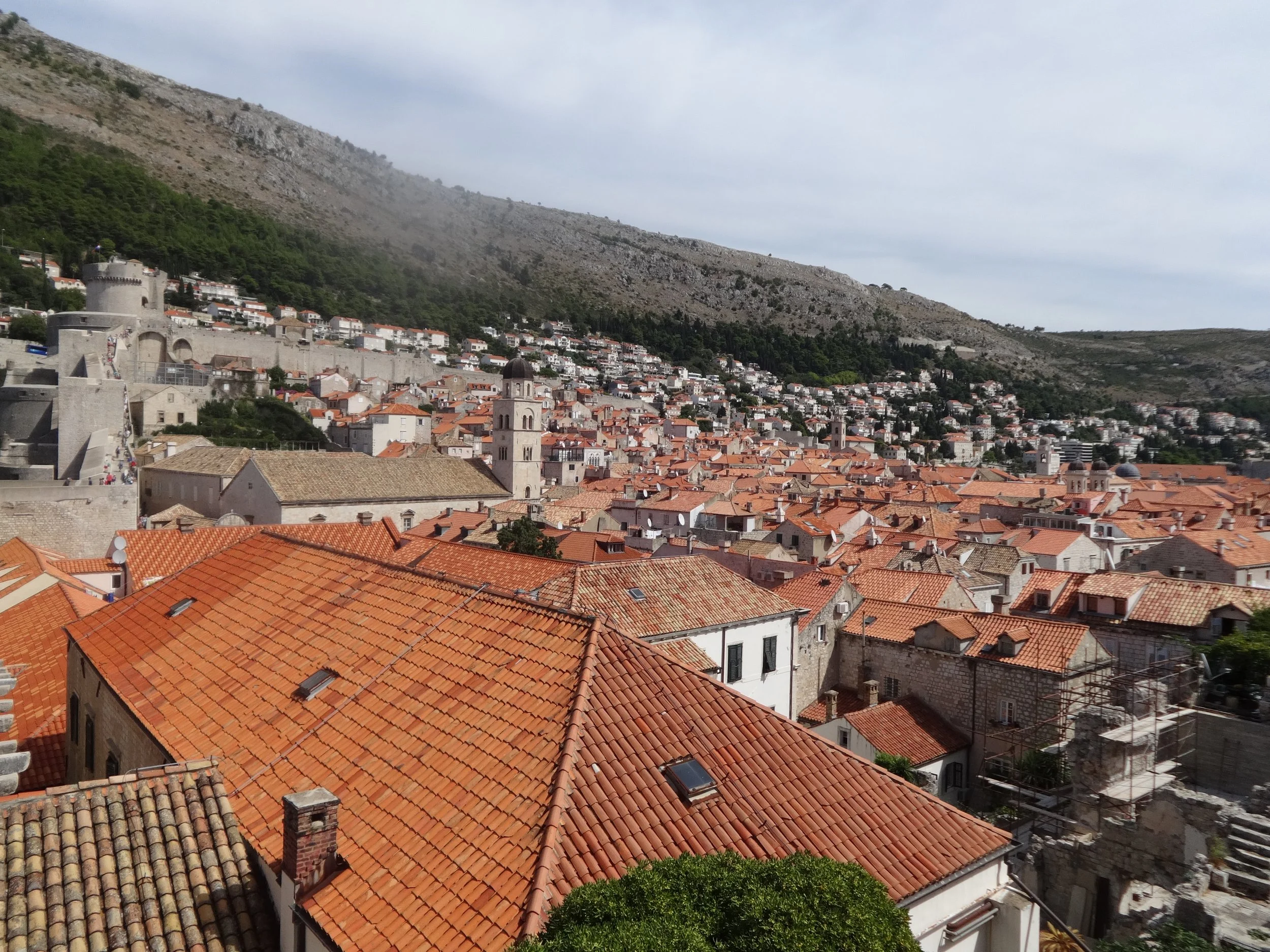 A view of a historic European city with red-tiled rooftops, old stone buildings, church towers, and a castle wall with a tower, set against a backdrop of green hills and a mountain.
