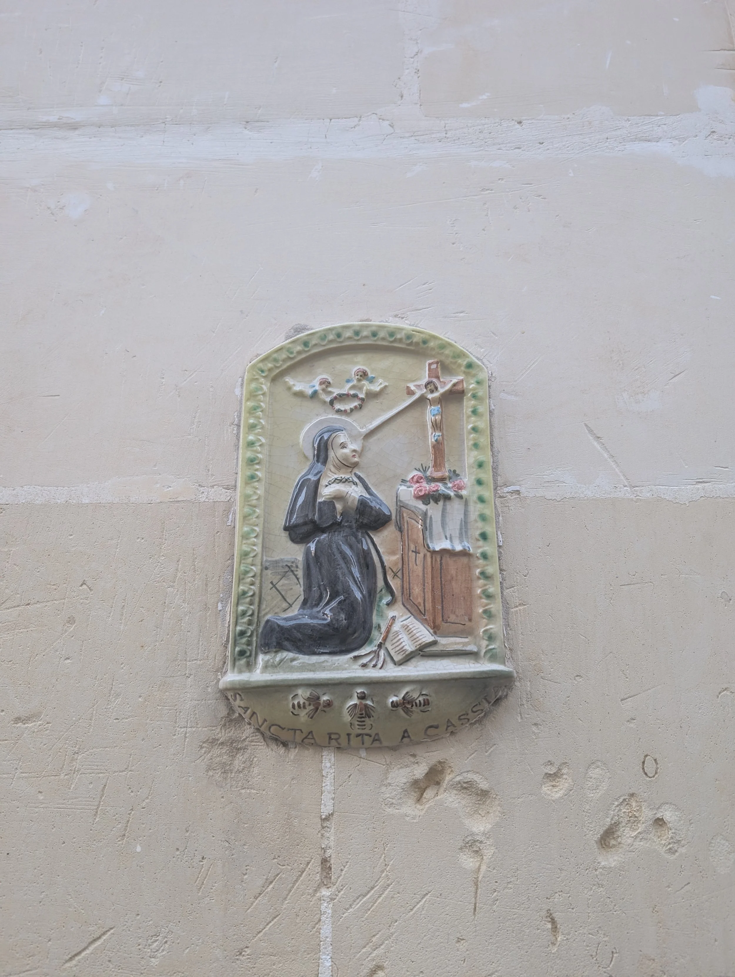 Ceramic wall plaque depicting a nun kneeling and praying, with a cross and religious symbols, mounted on a beige wall with inscriptions around the border.